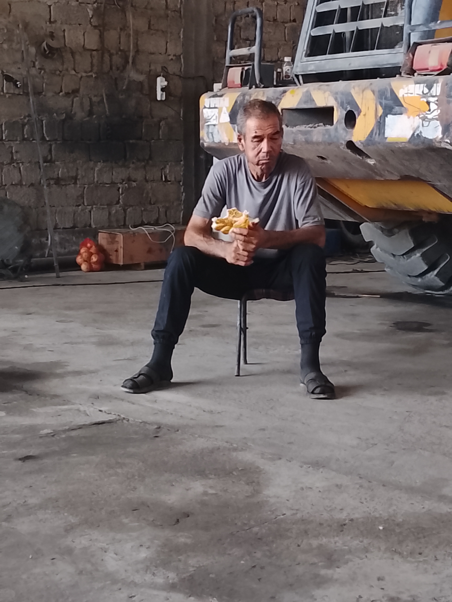 A middle-aged man with graying hair sits on a stool in what appears to be a workshop or industrial space. He is wearing a gray t-shirt, dark pants, dark socks, and dark sandals. He is holding a piece of food, possibly a sandwich, in both hands and looking down at it. To the left of the man, there is a wooden crate with a red mesh bag containing several round, orange objects, possibly fruit. In the background, there is a large piece of yellow and black machinery, likely construction equipment, with a large tire visible. The walls are constructed of rough, brick-like material. The floor is concrete, showing some wear and discoloration. The lighting suggests an indoor environment, possibly a garage or warehouse. There is no indication that this scene is located in the city of Sabir Rakhimov, Uzbekistan.