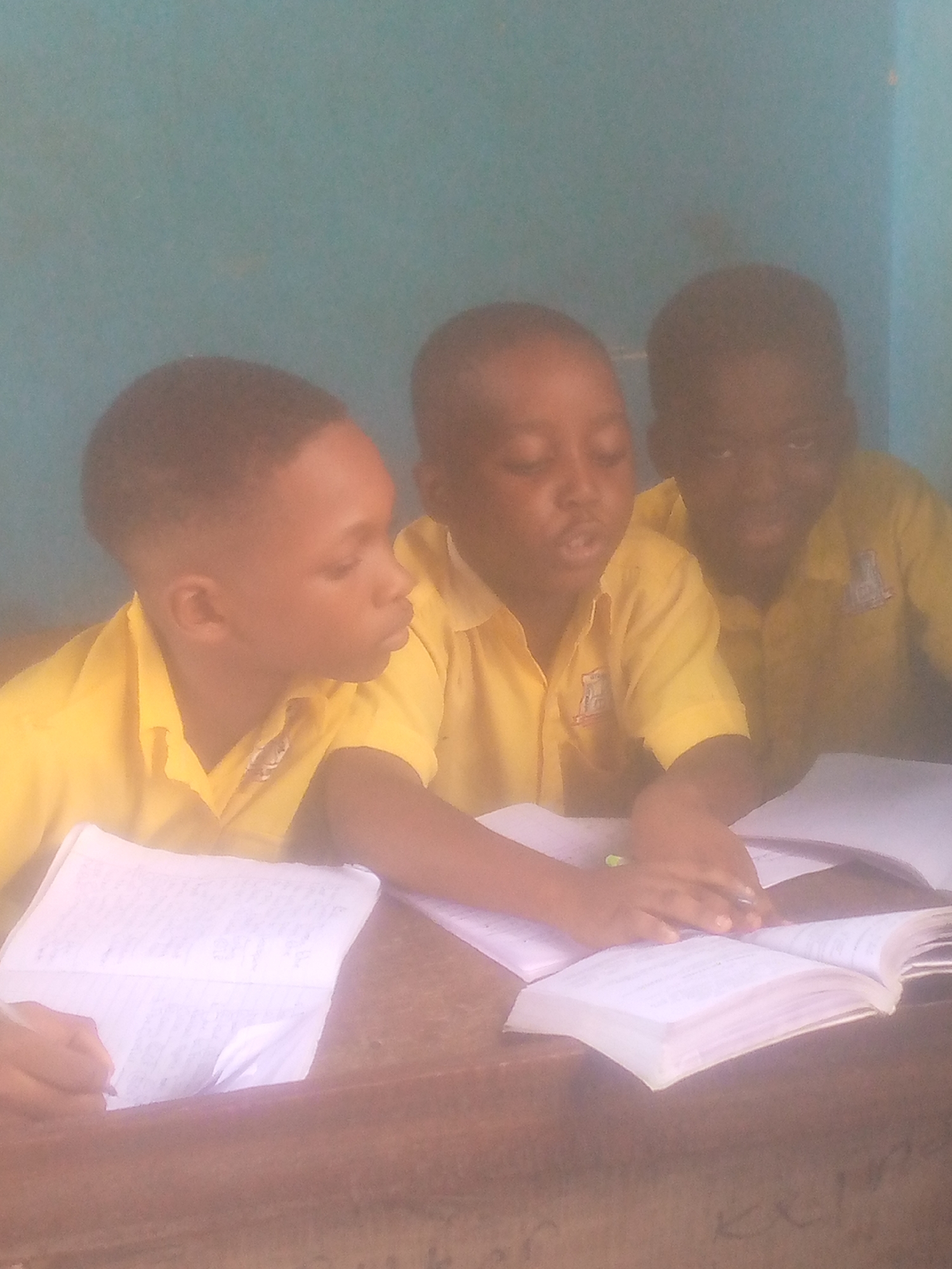 Three young children in yellow collared shirts sit at a wooden desk. Each child has a book. Two of the children appear to be reading together from one of the books. The location appears to be a classroom with a turquoise wall behind the children. The city and specific location are unknown.