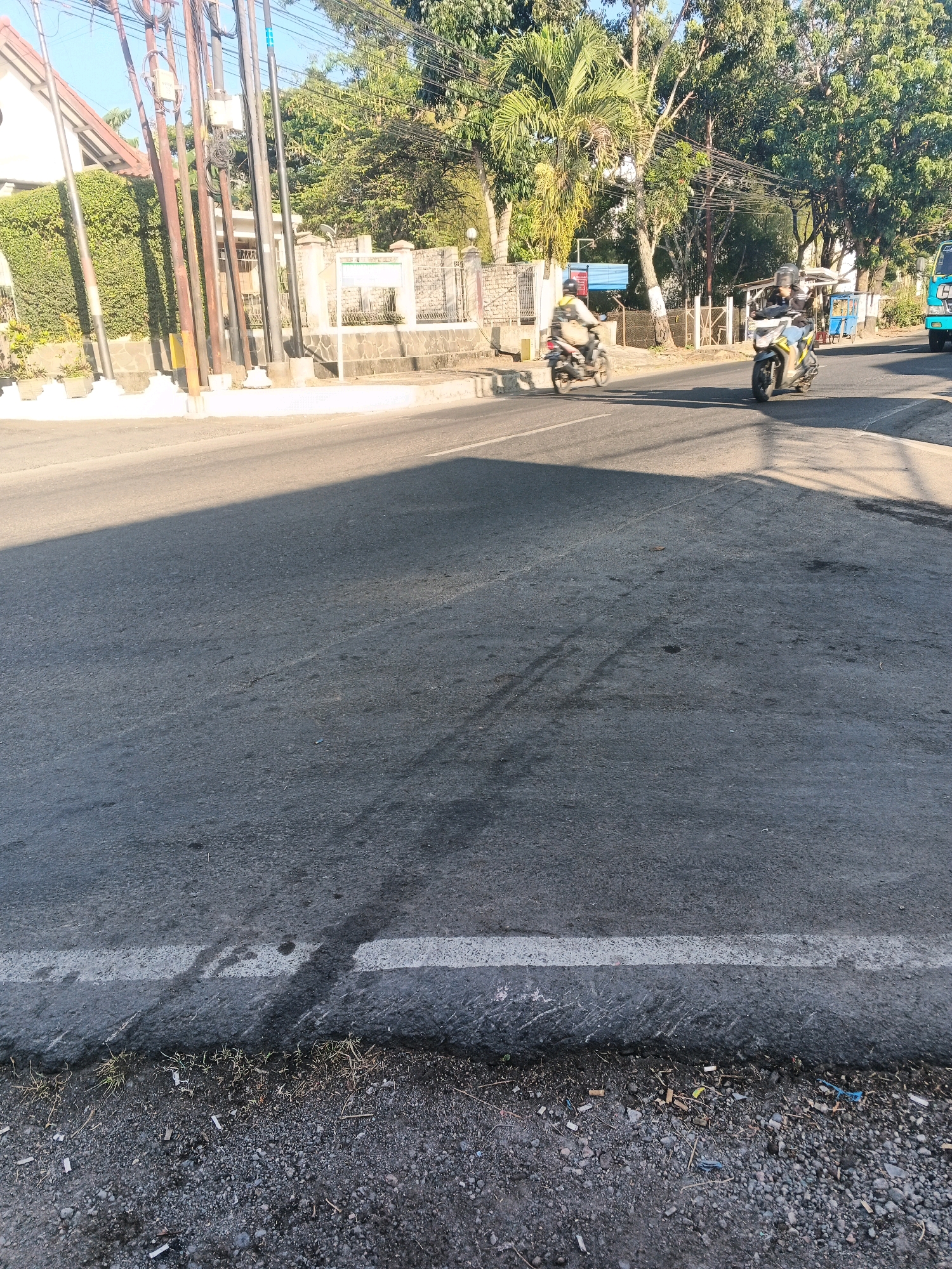 The image depicts a street scene in Semarang, Indonesia, during daylight. The road surface is asphalt with a white line marking. Shadows from overhead structures, likely utility poles and trees, cast long diagonal shapes across the road. On the left side of the road, a white building with a tiled roof and green foliage is visible, flanked by several utility poles. In the background, trees with dense green foliage form a backdrop. Two motorcycles are in motion on the road. The motorcycle in the foreground is yellow and white, ridden by an individual wearing a helmet and a light-colored jacket. Further down the road, another motorcycle, appearing lighter in color, is also in motion with a rider. A blue truck is partially visible on the far right edge of the image. The ground beside the road is covered in gravel and sparse dry grass, with some cigarette butts scattered on it. The sky is clear and blue, suggesting fair weather.