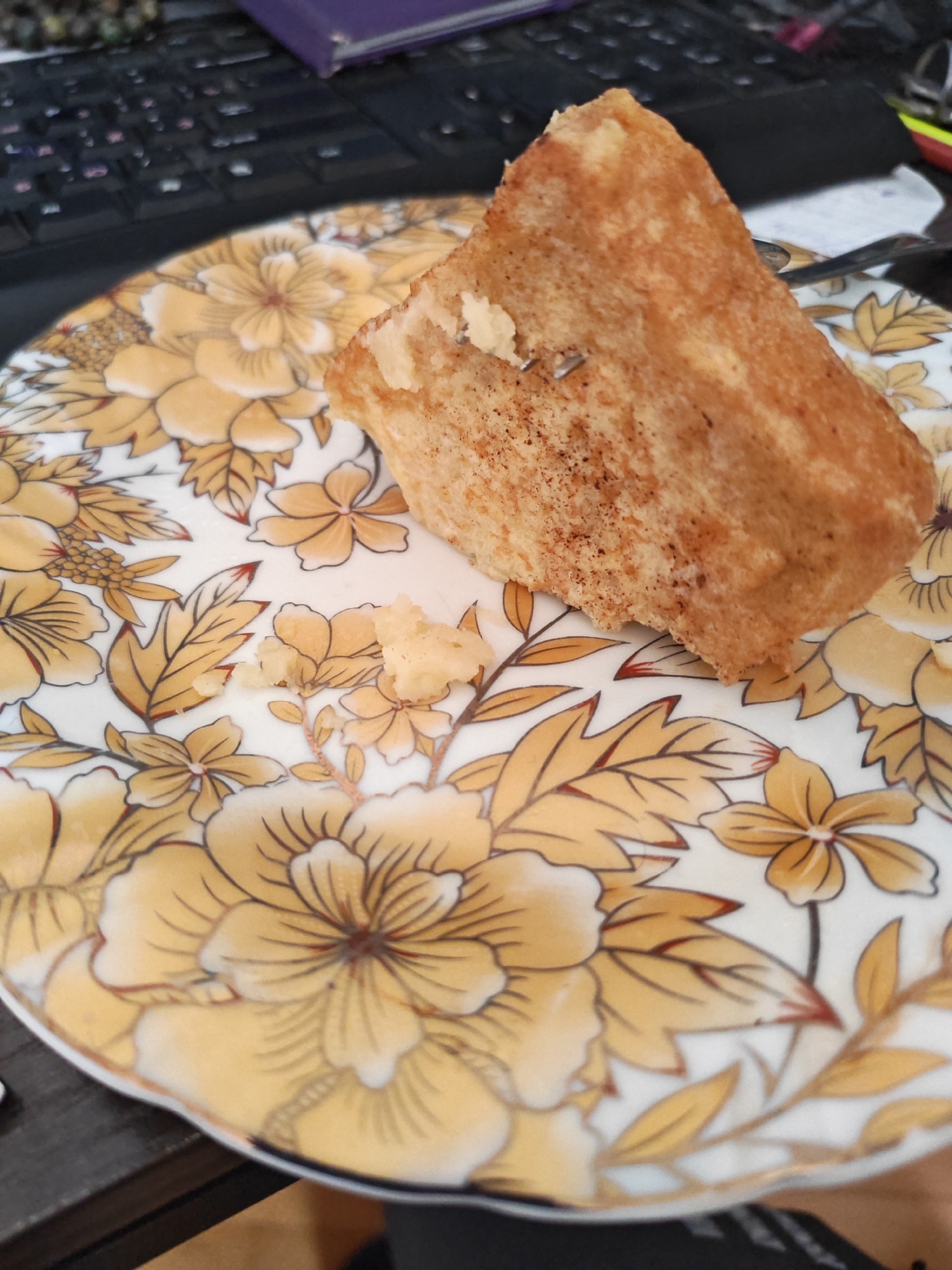 A close-up, overhead shot shows a piece of cake on a floral patterned plate. The cake is golden brown and appears to be made of sponge cake. It is cut into a triangular wedge and has a few crumbs scattered on the plate. The plate has a white background with yellow and brown floral designs. A fork is partially visible on the right side of the plate, its tines resting on the cake. In the background, a dark computer keyboard and a purple book are visible, suggesting the scene is likely indoors. The overall impression is a simple, everyday moment of enjoying a treat, possibly at a desk or table. The scene does not contain any information that would place it in Vanadzor, Armenia.
