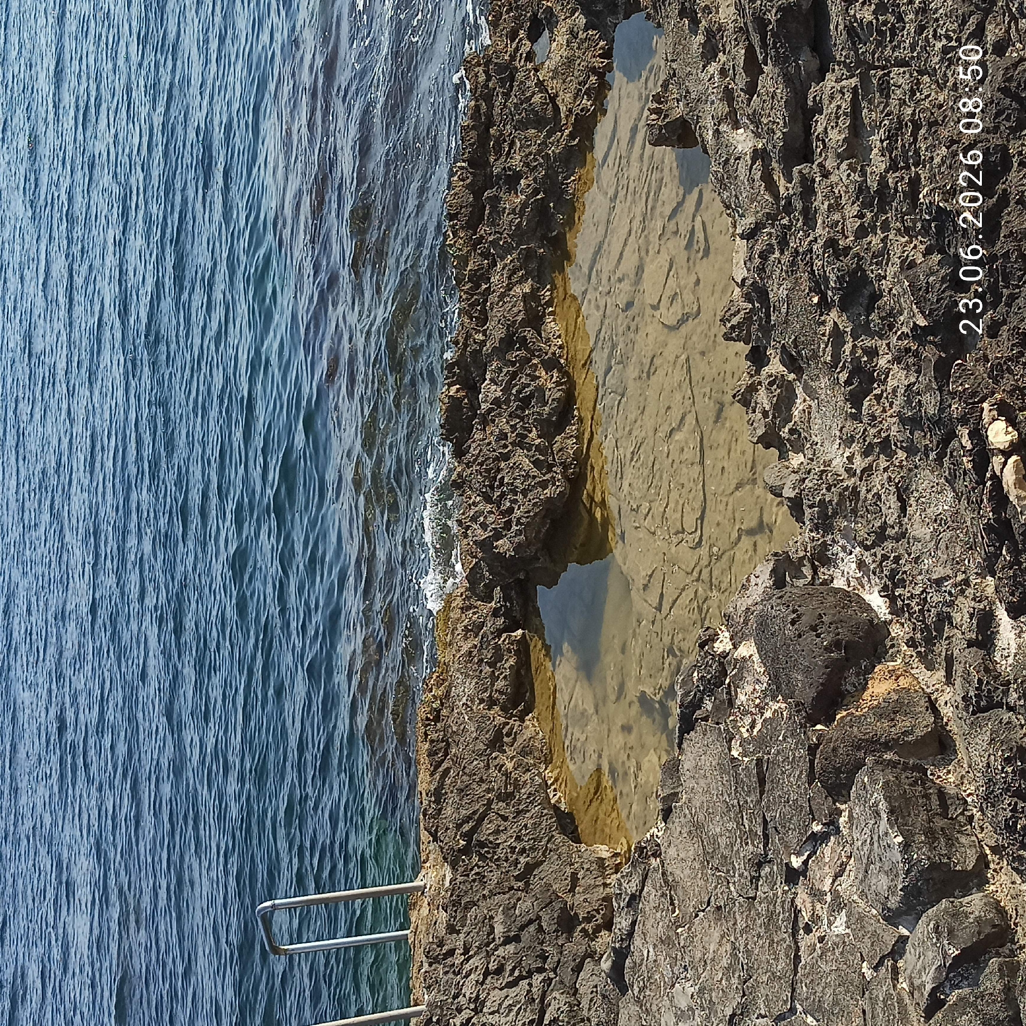 The image depicts a natural tidal pool formed within rough, dark volcanic rock formations along the coastline. The pool is filled with clear, shallow water, revealing the sandy and rocky bottom beneath. The sea, a deep blue, stretches into the background, with gentle waves lapping at the shore. On the left side of the frame, two metal railings of a staircase emerge from the rocks, suggesting a point of access to the water. The overall scene is natural and serene, likely located in a coastal area. A timestamp in the bottom right corner reads "23.06.2026 08:50".