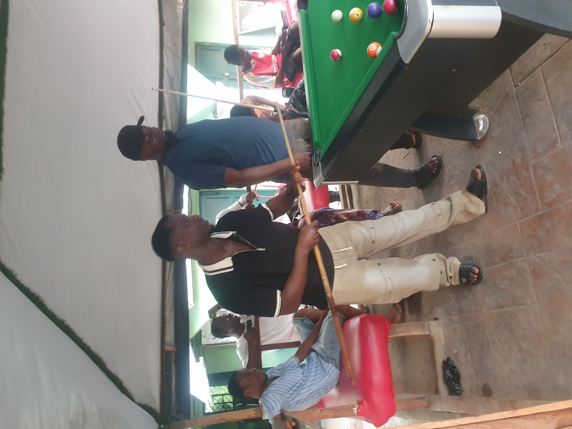 The image depicts a group of people engaged in a game of pool in an outdoor setting. Three visible players are actively involved, while others observe. The pool table is positioned on a tiled floor under a tent-like structure. There are pool balls on the table. The scene appears to be in a recreational area or potentially a public space in Lokoja, Nigeria. The environment includes details of the local architecture and community activities.