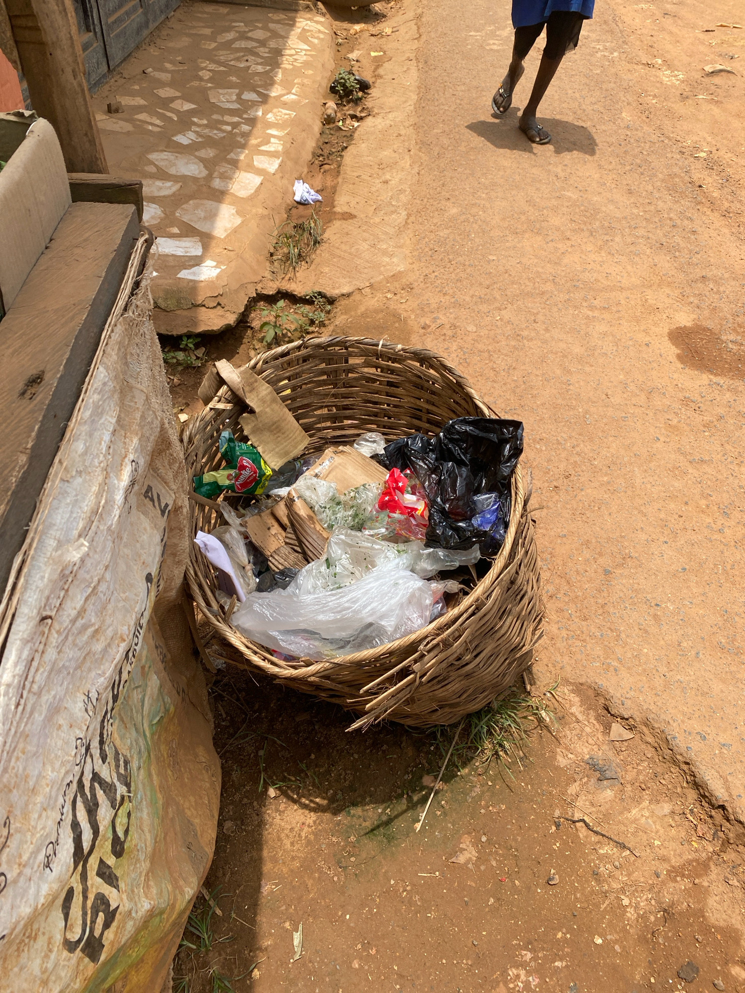 In Iworoko, Nigeria, a close-up shot captures a woven basket overflowing with refuse, positioned on a dusty, reddish-brown dirt surface adjacent to a paved walkway. The walkway, composed of irregular light-colored stones embedded in a dirt base, is partially shaded by the sun, creating a dappled effect. To the left of the basket, a large, weathered burlap sack leans against a wooden structure.
The basket itself contains a variety of discarded items, including crumpled plastic bags, some of which are black and others transparent, revealing plastic wrappers and what appear to be food scraps. Pieces of cardboard and green leafy material are also visible within the trash.
In the background, a person is walking away from the camera on the dirt road. They are wearing dark leggings, a blue garment, and dark sandals. Their legs and feet are visible, casting a shadow on the ground. The overall scene depicts an outdoor urban environment with elements of both everyday life and waste disposal.