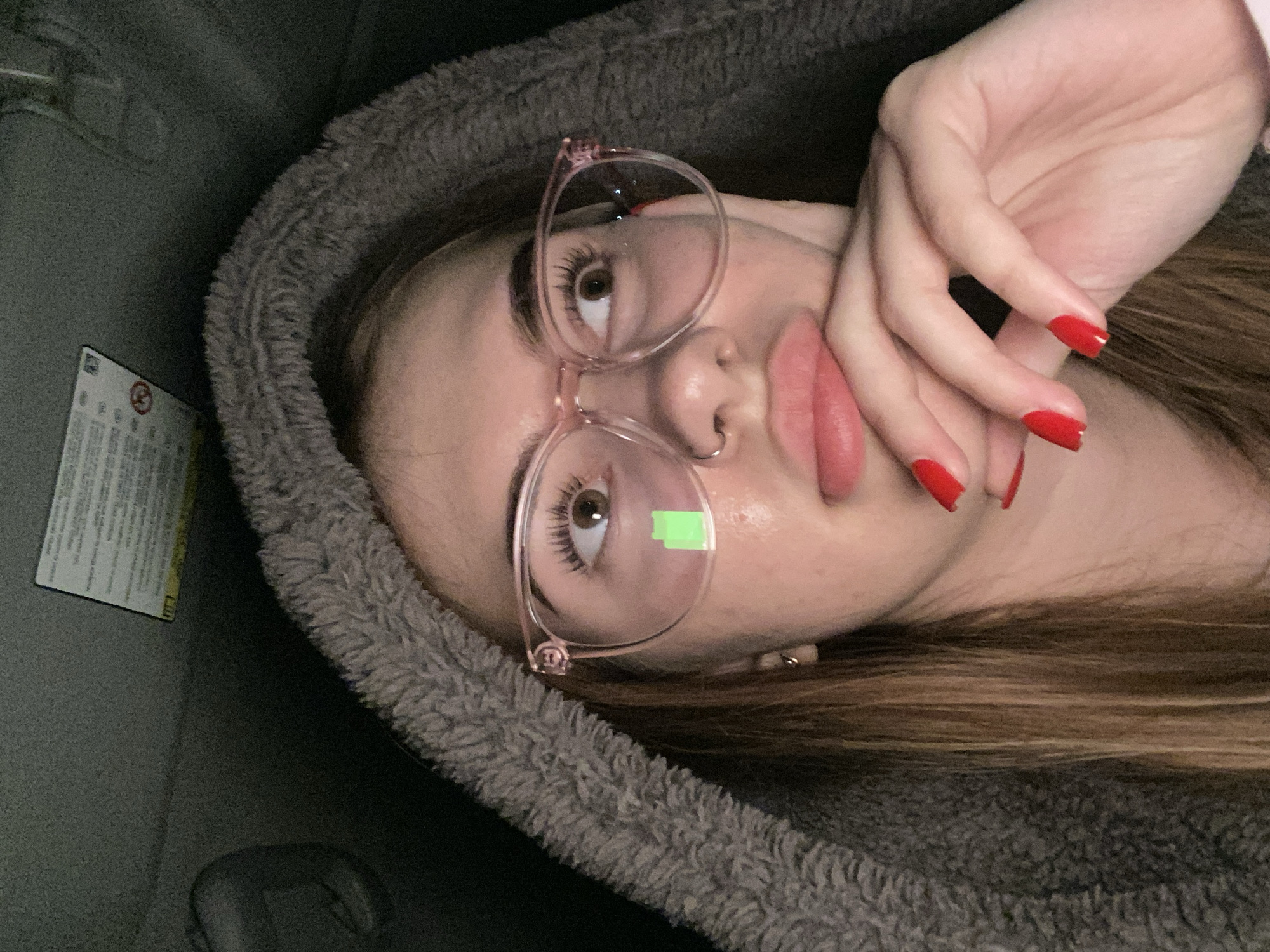 A portrait-style image, rotated 90 degrees clockwise, captures a young person from a close-up, slightly elevated perspective. The person is wearing clear-framed eyeglasses, with a small, bright green rectangular sticker or reflection visible on the right lens. They have dark, long eyelashes and are looking upwards and slightly to the right. Their lips are slightly parted in a pout, and a small ring is visible in their left nostril.
A hand, adorned with bright red nail polish, rests gently near their chin and mouth. The person's hair, a mix of light brown and auburn with subtle highlights, peeks out from under a dark gray, fuzzy hood or blanket that frames their face and shoulders.
The setting appears to be a dimly lit interior, strongly suggesting a vehicle. On the far left of the frame, a white rectangular label with black text is partially visible, typical of warning labels found on car sun visors or interior panels. While most of the text is illegible, a circled 'R' (registered trademark symbol) and a 'no' or 'X' symbol within a circle can be discerned on the label. The overall mood is casual and contemplative.