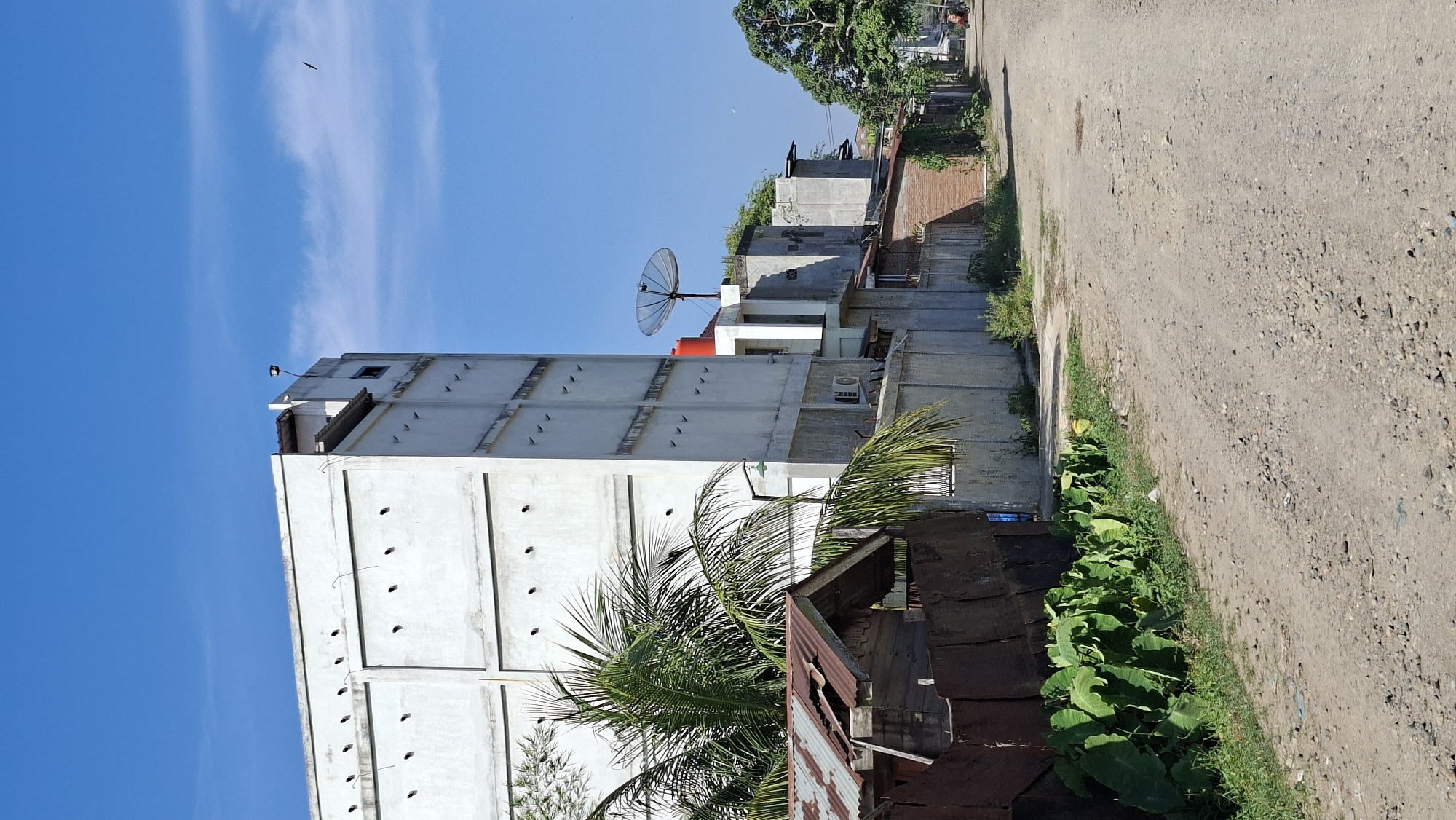 The image depicts a scene in the city of Keudebayu, Indonesia, on a clear day with a bright blue sky. A dirt road runs through the frame, flanked by buildings and vegetation.
On the left side of the road, a tall, white, multi-story building dominates the foreground. It has a series of small, dark, rectangular openings along its facade. In front of this building, a smaller, dilapidated structure with a corrugated tin roof is visible, partially obscured by lush green foliage. A palm tree with large fronds extends into the frame from the left.
Further down the road, to the right of the tall white building, are other structures. One building has a red water tank on its roof and a satellite dish. Another building is constructed with brick. Green vegetation lines the sides of the road, and trees with dense foliage are present further in the distance.
The road itself is unpaved and appears to be made of dirt and gravel, with some scattered stones. In the far distance, a person on a motorcycle is visible, suggesting ongoing activity in the area. The overall environment appears to be a mix of residential and possibly commercial or agricultural structures, characteristic of a developing urban or semi-urban area. The lighting suggests it is daytime, with the sun casting shadows.
