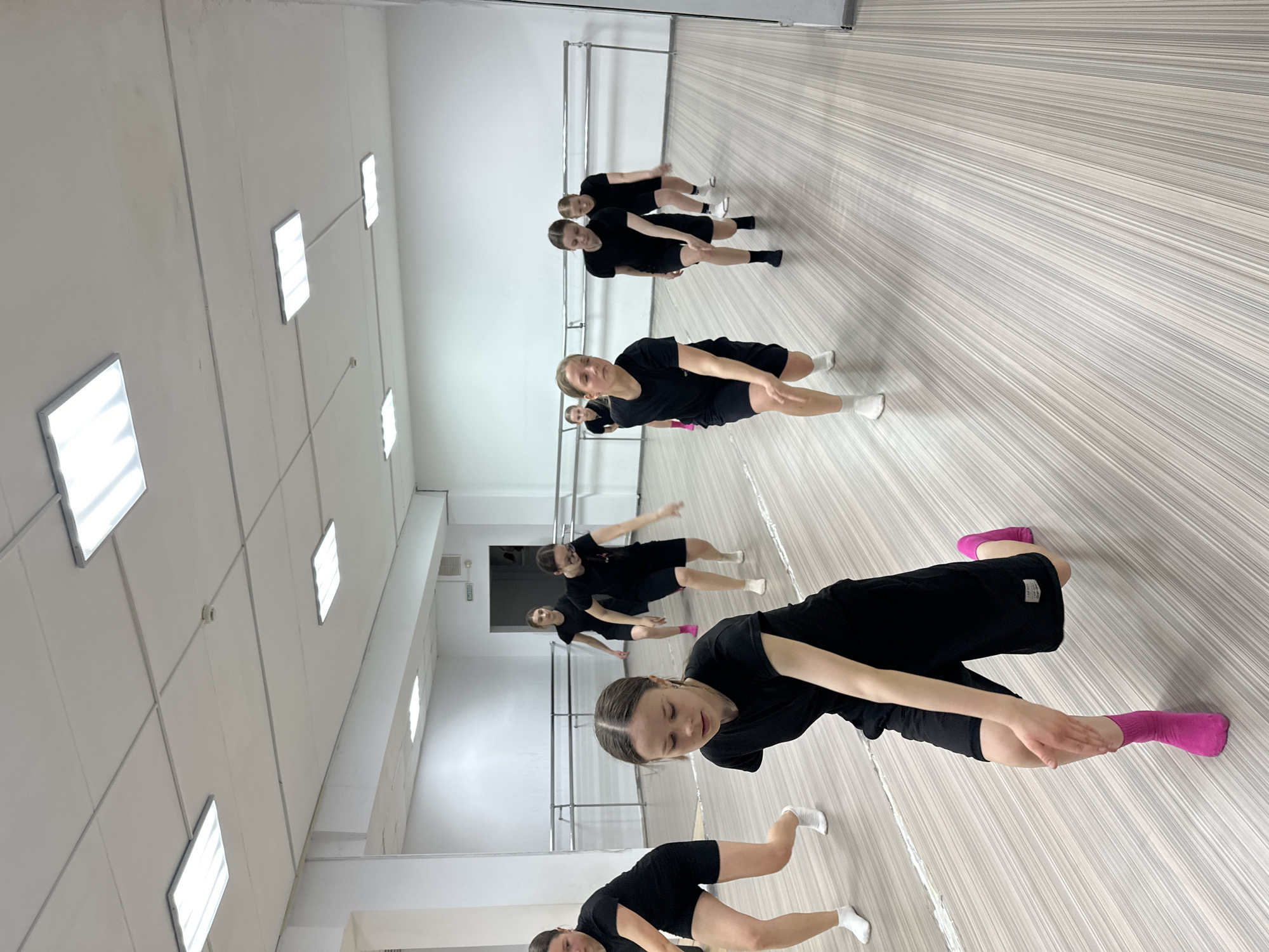 A group of approximately seven young individuals, predominantly female, are engaged in a dance or exercise class within a brightly lit indoor studio in Votkinsk, Russia. They are uniformly dressed in black athletic t-shirts and shorts. Most wear white socks, though one person in the mid-foreground is distinguishable by bright pink socks. The participants are performing a synchronized movement, leaning forward at the waist with bent knees, appearing focused on their posture or stretch.
The studio features plain light-colored walls with multiple horizontal metal dance barres running along one side. The floor exhibits a distinct light brown and grey striped wooden or laminate pattern that contributes to the room's perspective. Overhead, the ceiling is fitted with several rectangular fluorescent light fixtures, ensuring ample illumination. In the background, near a dark doorway, a white sign with black Cyrillic text reads "ВЫХОД" (Vykhod), meaning "Exit" in Russian. The scene depicts a focused and active practice session.
