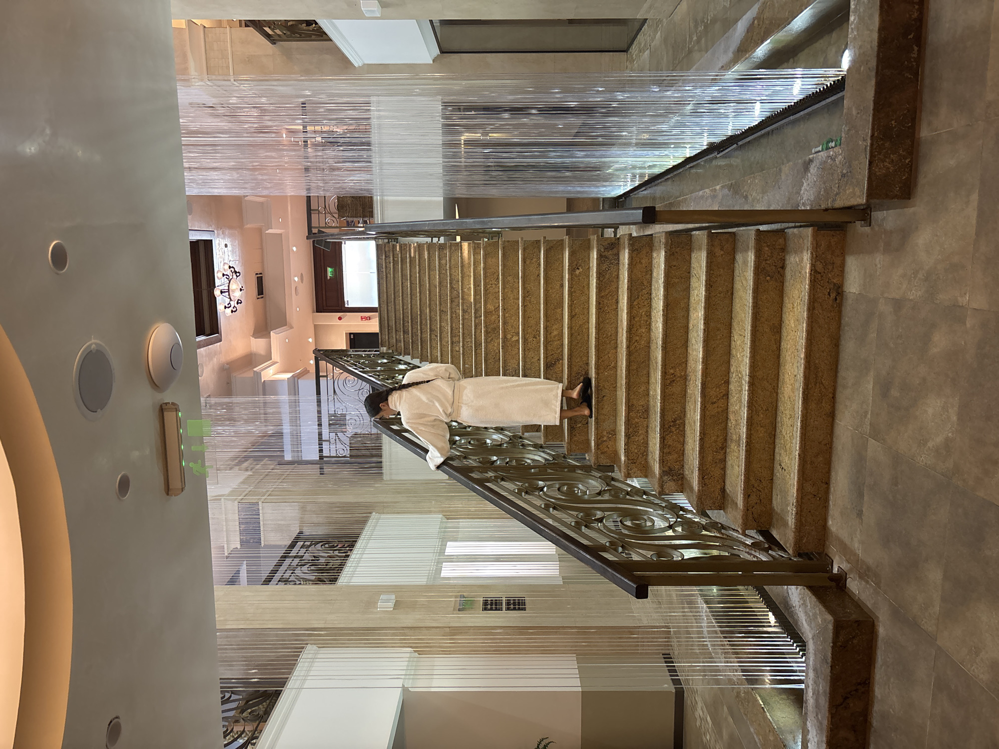 A person wearing a white bathrobe walks up a wide, marble staircase in an indoor atrium. The staircase is flanked by ornate metal railings and a translucent water feature composed of vertical strands of light. The atrium has a high ceiling with recessed lighting and decorative architectural elements. To the left of the stairs, a white wall with a window is visible, and on the right, a hallway with glass doors and more water features can be seen. The floor is tiled in a neutral tone. There are "EXIT" signs visible above a doorway. The overall impression is one of a luxurious spa or hotel interior. The location is identified as Razliv, Bulgaria.