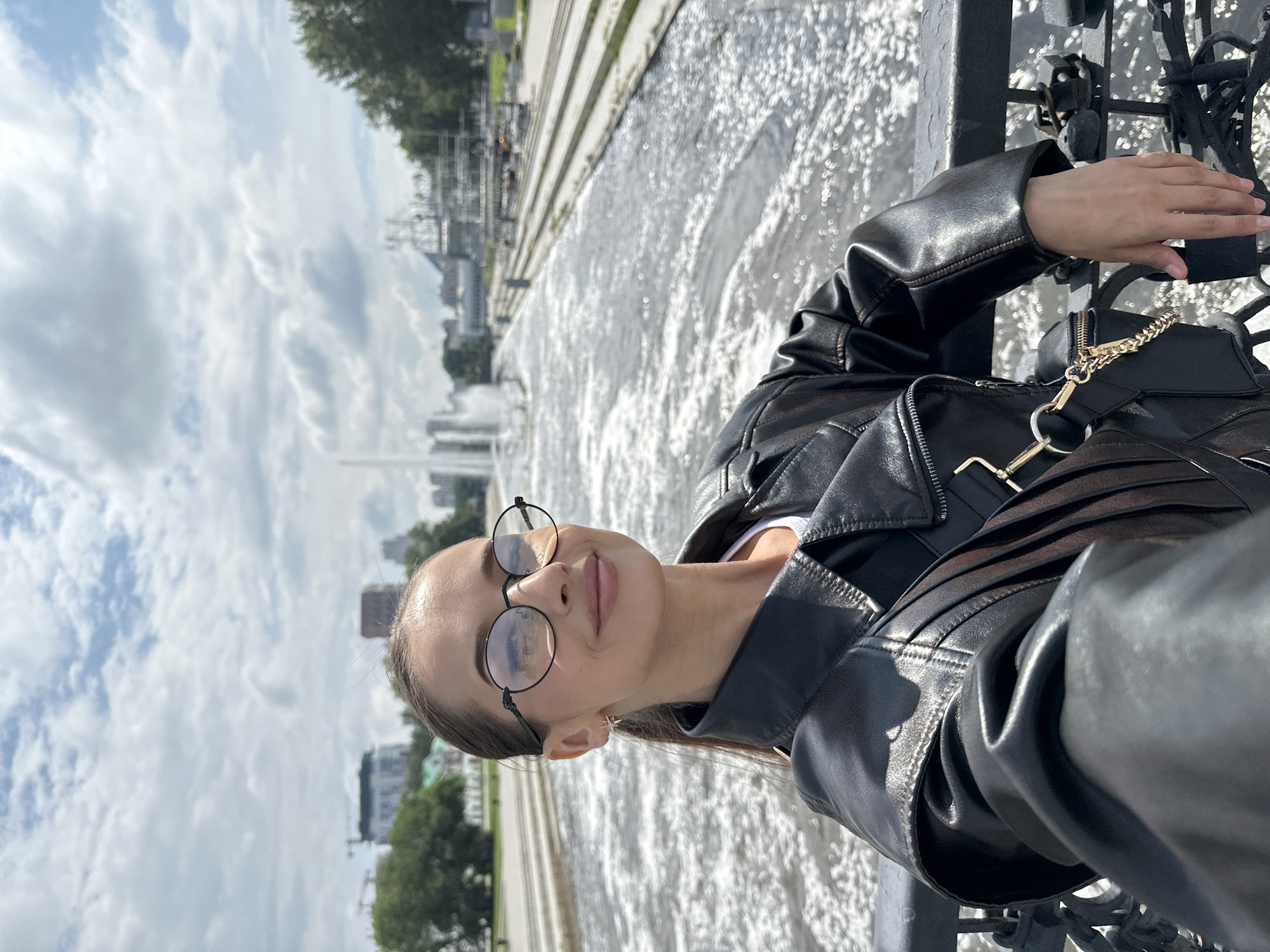 A young woman wearing round glasses and a black leather jacket takes a selfie on a bridge overlooking a wide, flowing canal. The canal is lined with concrete embankments and in the background, a large fountain sprays water into the air. Beyond the fountain, the city skyline of Yekaterinburg, Russia, is visible, featuring buildings and trees. The sky is partly cloudy. The woman is leaning against a black railing adorned with numerous padlocks, a common sight on bridges in Yekaterinburg, signifying promises and love.