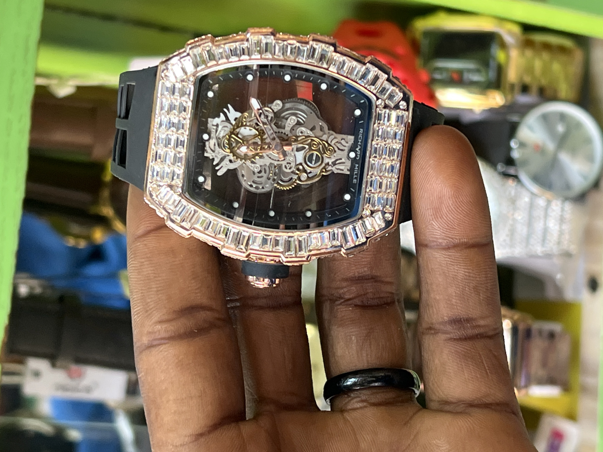 A close-up, shallow depth-of-field shot shows a person's hand holding a luxury watch. The watch has a tonneau-shaped rose gold case encrusted with baguette-cut diamonds. The dial is black and features a visible, intricate mechanical movement with gold-toned gears and bridges. The watch has a black rubber strap. The person is wearing a black ring on their left ring finger. The background is blurred and appears to be a retail environment, possibly a market stall in Bukuru, Nigeria, with shelves containing other items, including what look like more watches and brightly colored goods. The lighting suggests an indoor setting, possibly with artificial light.