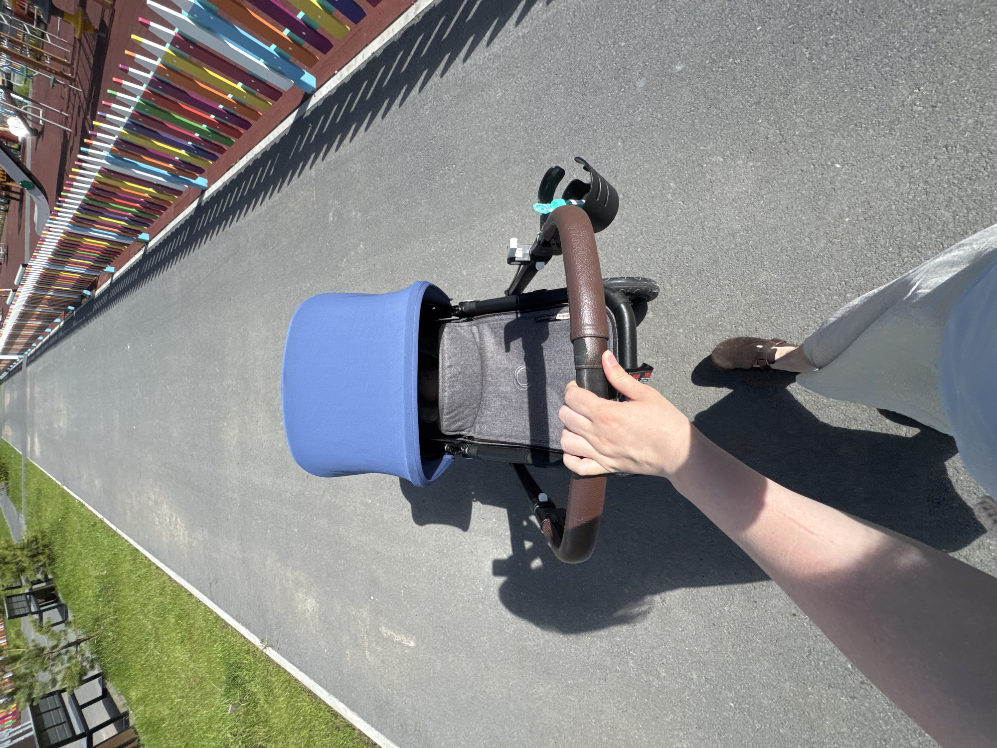 The image shows a person's arm and hand pushing a stroller on a paved path next to a grassy area. The stroller has a blue canopy and a brown leather handle. A black cup holder is attached to the stroller handle. In the background, there is a colorful fence with a shadow cast by it. The sun is shining, creating shadows. The scene appears to be outdoors in a park or recreational area. The time of day is likely daytime due to the bright sunlight and strong shadows. The location context provided is Patrusheva, Russia.