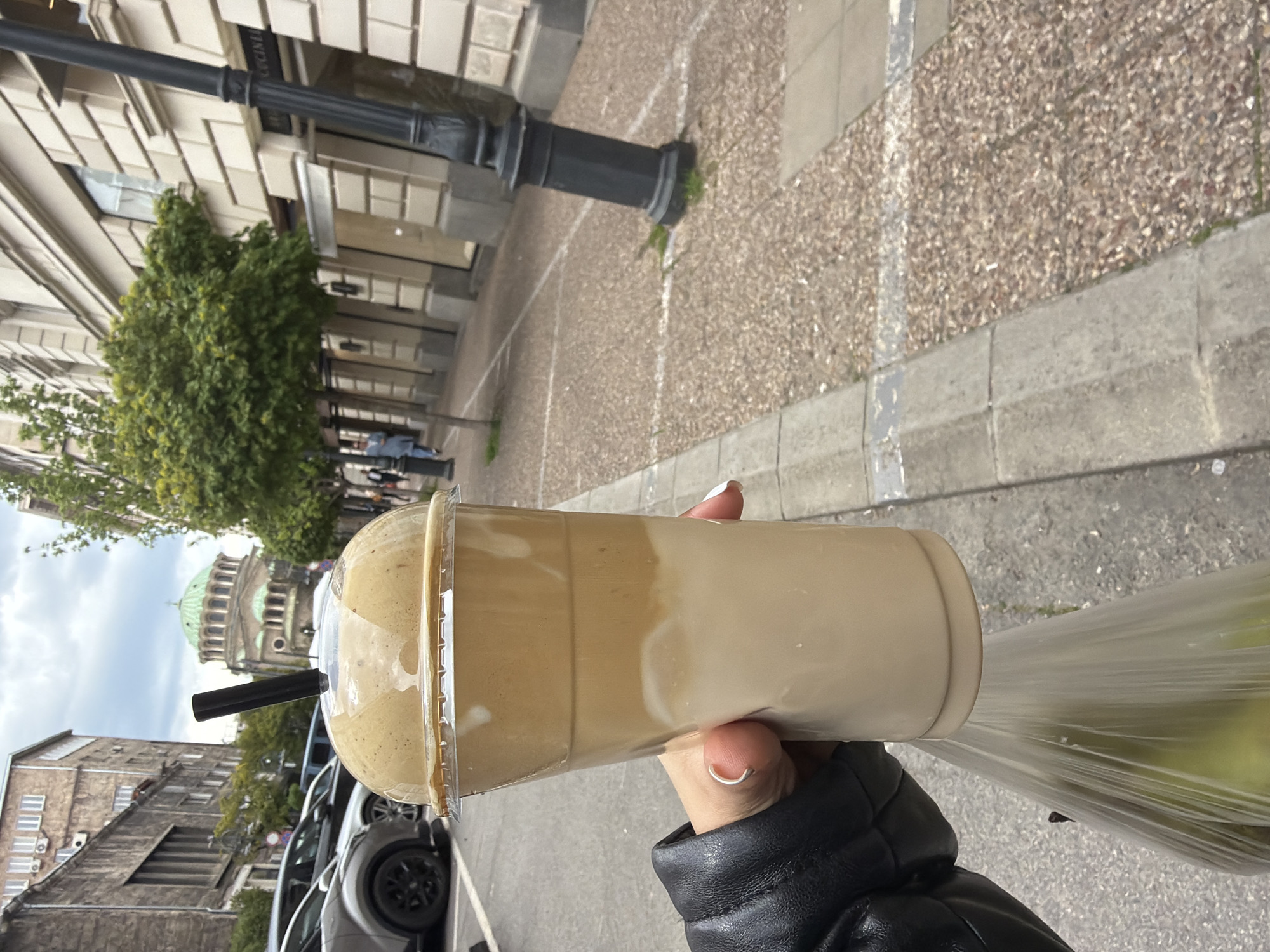 A person holds a large plastic cup of iced coffee with a black straw in front of a street scene. The cup is slightly tilted, revealing a two-tone coffee mixture. The person wears a black leather jacket and a ring. Behind the cup, a street stretches into the distance, lined with parked cars and buildings. A large domed building, likely a cathedral, is visible among the trees. The environment suggests an urban setting, likely a city street. Given the architectural style and presence of the Alexander Nevsky Cathedral, the image location is Sofia, Bulgaria.