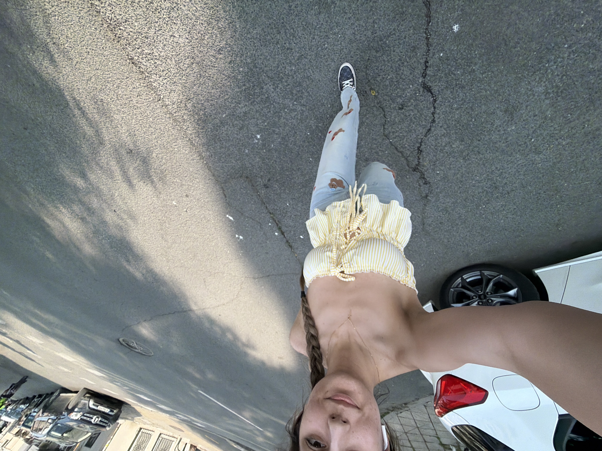 This image presents an overhead, angled view of a young woman standing on an asphalt street in Burgas, Bulgaria. She is looking downwards towards the camera with a neutral expression. Her long, braided brown hair falls over her shoulder. She is dressed in a yellow and white striped ruffled crop top, light blue ripped jeans with brown patches, and black sneakers. A delicate gold body chain is visible on her torso, and she wears white wireless earbuds.
The street surface is asphalt, showing various textures and minor cracks. Strong shadows, likely cast by trees or buildings, stretch across the road, indicating clear, sunny daytime conditions. Parked vehicles are visible on both sides of the street: a white car with a prominent red taillight and part of a wheel is on the right, and several darker vehicles are further down the street on the left. Buildings line the background on the left side of the street. A manhole cover is partially visible on the asphalt to the left. No legible text is discernible within the image.