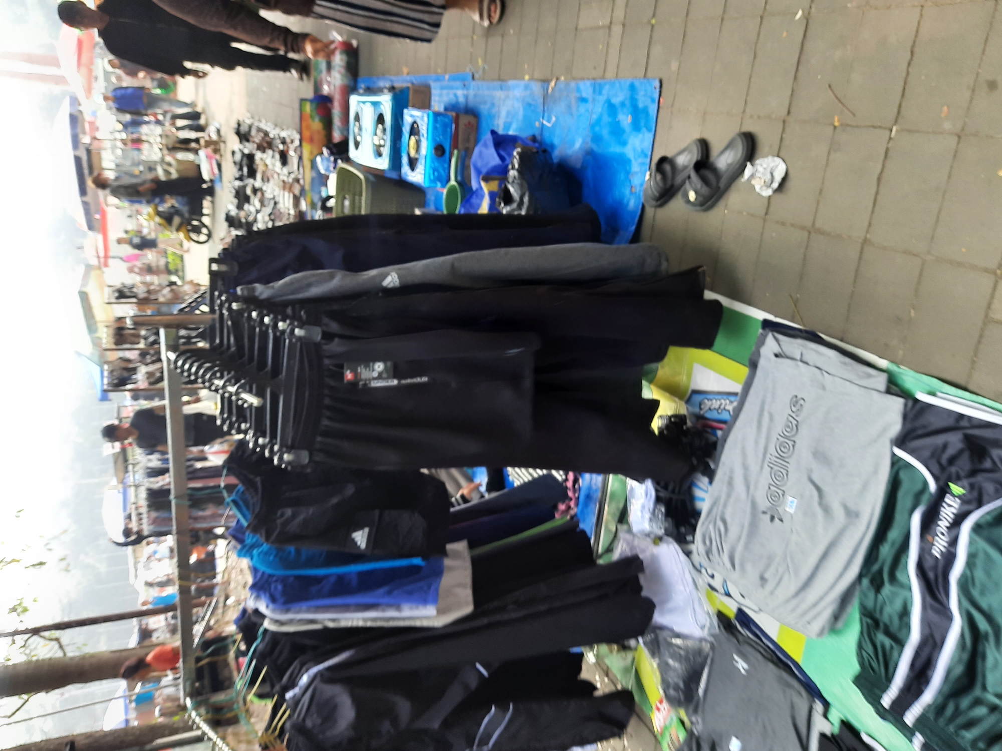 A street market scene is depicted in Bandung City, Indonesia. The foreground shows several racks of clothing, primarily black and grey tracksuits and shorts. Some garments have visible brand logos, including "adidas" and "PRONIK". A blue tarp is spread on the ground to the right, displaying several portable gas stoves and other items. In the background, a number of people are browsing the stalls. A motorcycle is parked amongst the crowd. The market appears to be outdoors, with trees and some structures visible in the distance, suggesting a bustling commercial area. The lighting is bright, indicating it is daytime.