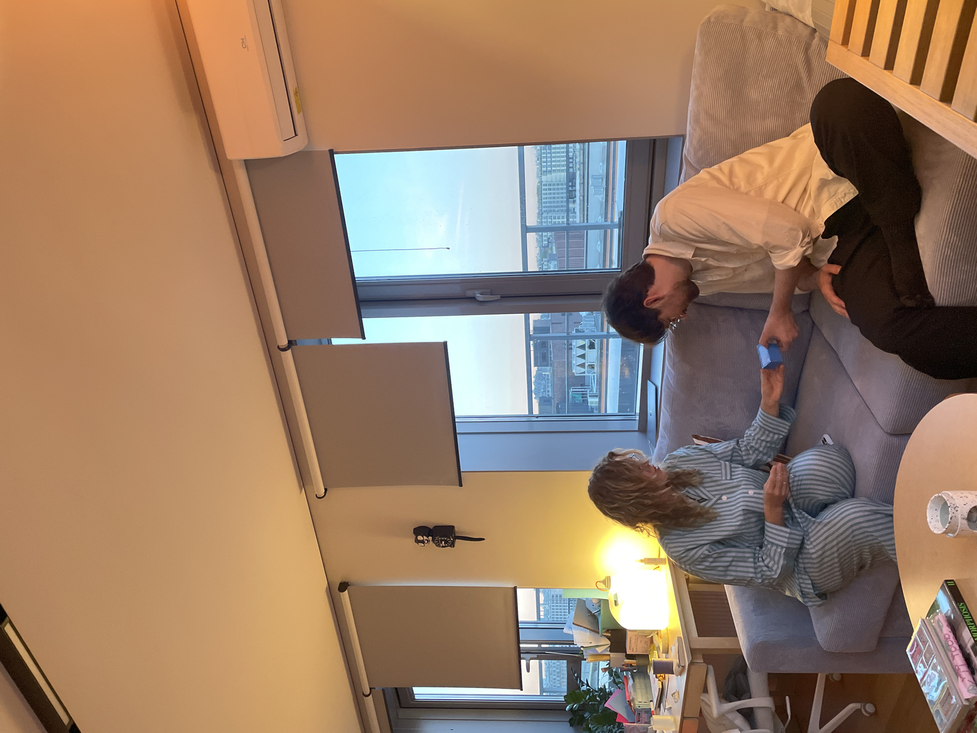 The image depicts an interior scene in what appears to be an apartment. A woman with blonde, curly hair, wearing striped pajamas, is seated on a light grey, corduroy-like couch. She is holding a small blue box. A man, wearing a light-colored shirt and dark pants, is standing next to the couch, also looking at the blue box. A round, light-colored table with a cup and books is beside the couch. A window in the background shows a city skyline with buildings. Three partially drawn roller blinds are visible on the window. A desk with a lamp, books, and various objects is visible beneath the window. Based on the architectural style and the urban environment, it is plausible that this scene is set in Saint Petersburg, Russia.