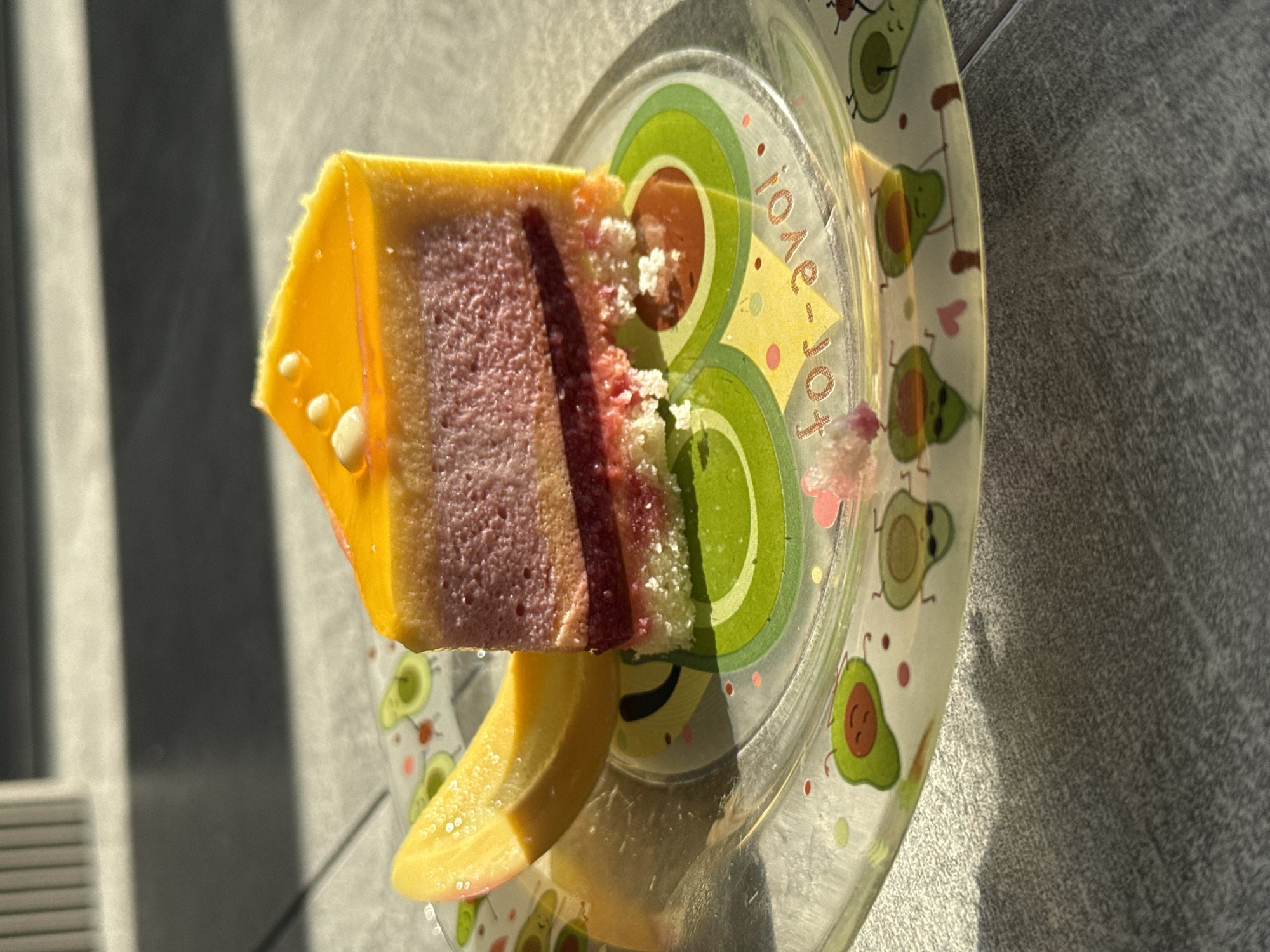 A slice of layered dessert with yellow glaze is presented on a clear plastic plate decorated with avocados and text. The plate is set on a gray textured surface under bright sunlight, possibly indoors near a window as indicated by the shadows. The exact location is Tomilino, Russia.