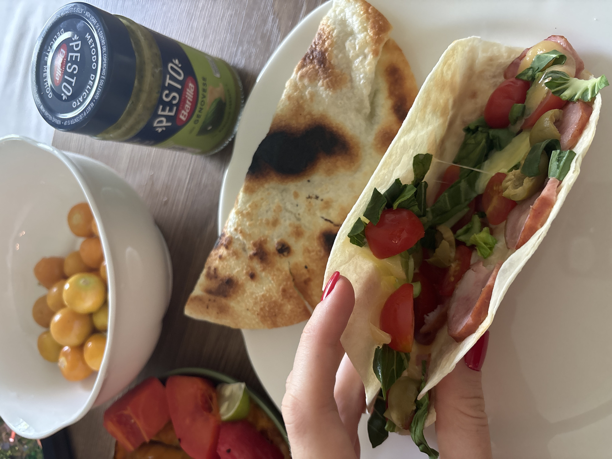 This close-up, overhead photograph captures a casual meal laid out on a light wooden surface, likely indoors in Choeng Thale, Thailand. The central focus is a hand with red nail polish holding a soft taco or wrap, bursting with fresh ingredients. Inside the wrap, visible fillings include halved red cherry tomatoes, green leafy vegetables, slices of cooked sausage or meat, melted cheese, and green olives.
On a white plate beside the held wrap, there's a large, irregularly shaped piece of golden-brown flatbread or calzone, exhibiting some darker, charred areas from cooking. To the left, a white bowl is partially visible, filled with a collection of small, round, yellow cherry tomatoes. Below this bowl, another plate displays cut red fruit, possibly watermelon or papaya, accompanied by a fresh lime wedge.
At the top left of the frame, a jar of Barilla Pesto Genovese is clearly visible. The label prominently features "Barilla PESTO GENOVESE," "METODO DELICATO," and "DELICATE METHOD," along with textual warnings in various languages regarding the lid's seal. The overall scene conveys a fresh and colorful eating experience.