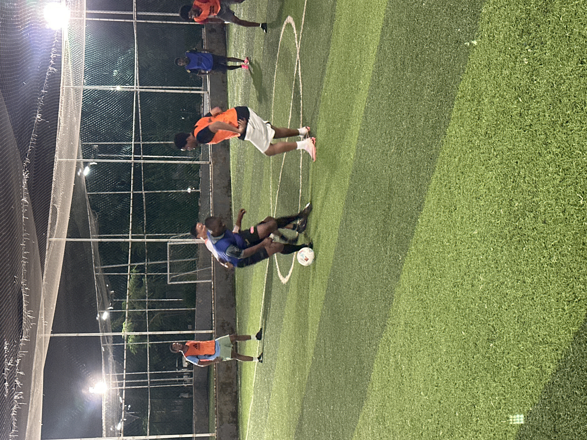The image depicts an outdoor, artificial turf soccer field at night, illuminated by artificial lighting. The scene is set in Abuja, Nigeria. Several men are engaged in a soccer match. One man in the foreground, wearing an orange bib over a blue shirt and light green shorts, is standing. To his right, another man in a blue shirt and black shorts is actively playing soccer, with a soccer ball at his feet. To the right of him, a third man in an orange bib and white shorts is also playing, facing away from the camera. In the background, two other men are visible, one in a blue bib and shorts, and another in an orange bib and dark shorts. The field is enclosed by fencing and netting, with trees and a building visible in the background beyond the fence.