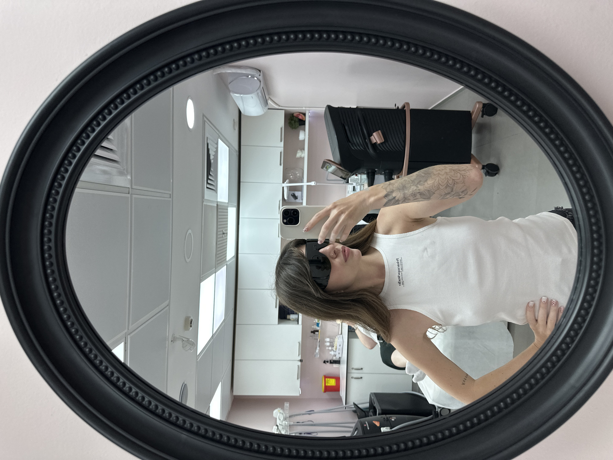 A person with a tattoo on their right forearm is visible in an oval, black-framed mirror. The person is wearing dark sunglasses and a white tank top with text on it. They are holding up a smartphone with their left hand to take a picture in the mirror. The mirror reflects a room with white tiled ceiling panels, white cabinets, and pink walls. A black medical chair with a pink strap is visible in the reflection. The location is in Tel-Aviv, Israel.