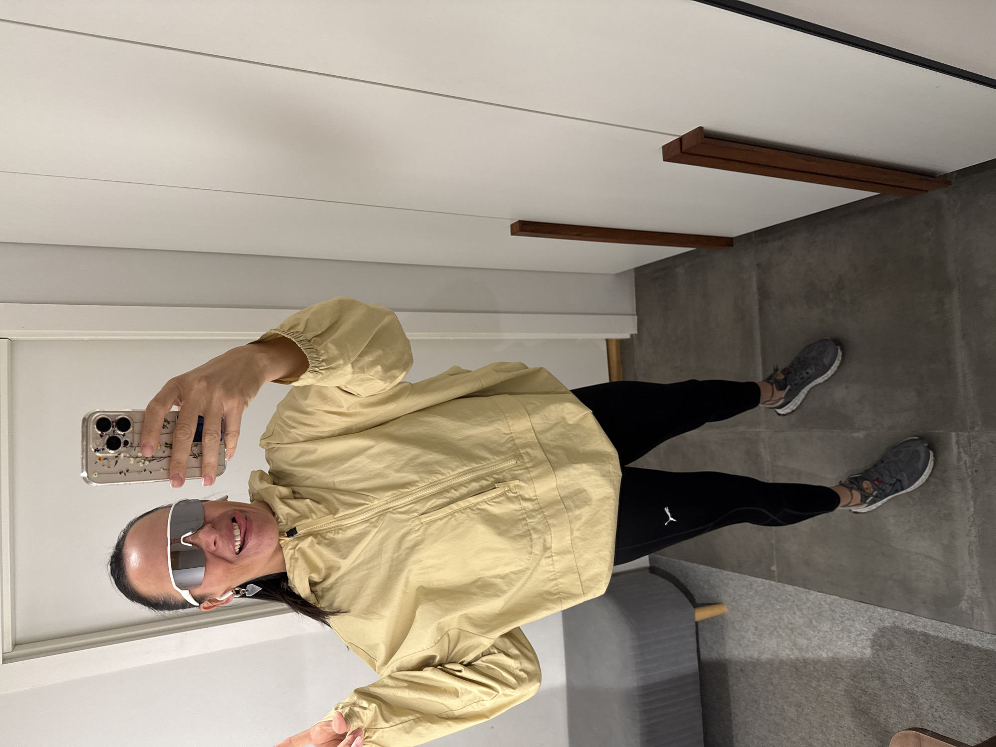 A woman stands in what appears to be a dressing room or a home with a modern aesthetic. She is taking a selfie with her phone. She is wearing large, white-rimmed sunglasses, a pale yellow hooded jacket with a quarter-zip, and black leggings with the Puma logo visible on the left leg. She also has on grey athletic shoes. Her tongue is sticking out slightly, and she is wearing a heart-shaped earring. The background consists of plain, off-white walls with vertical paneling and a dark grey floor. To her left, a portion of a grey bench with wooden legs is visible. To her right, there are two vertical wooden planks leaning against the wall. The lighting is bright and even, suggesting an indoor setting. There is no indication of the city of Chaikovsky, Russia, or any other specific location.
