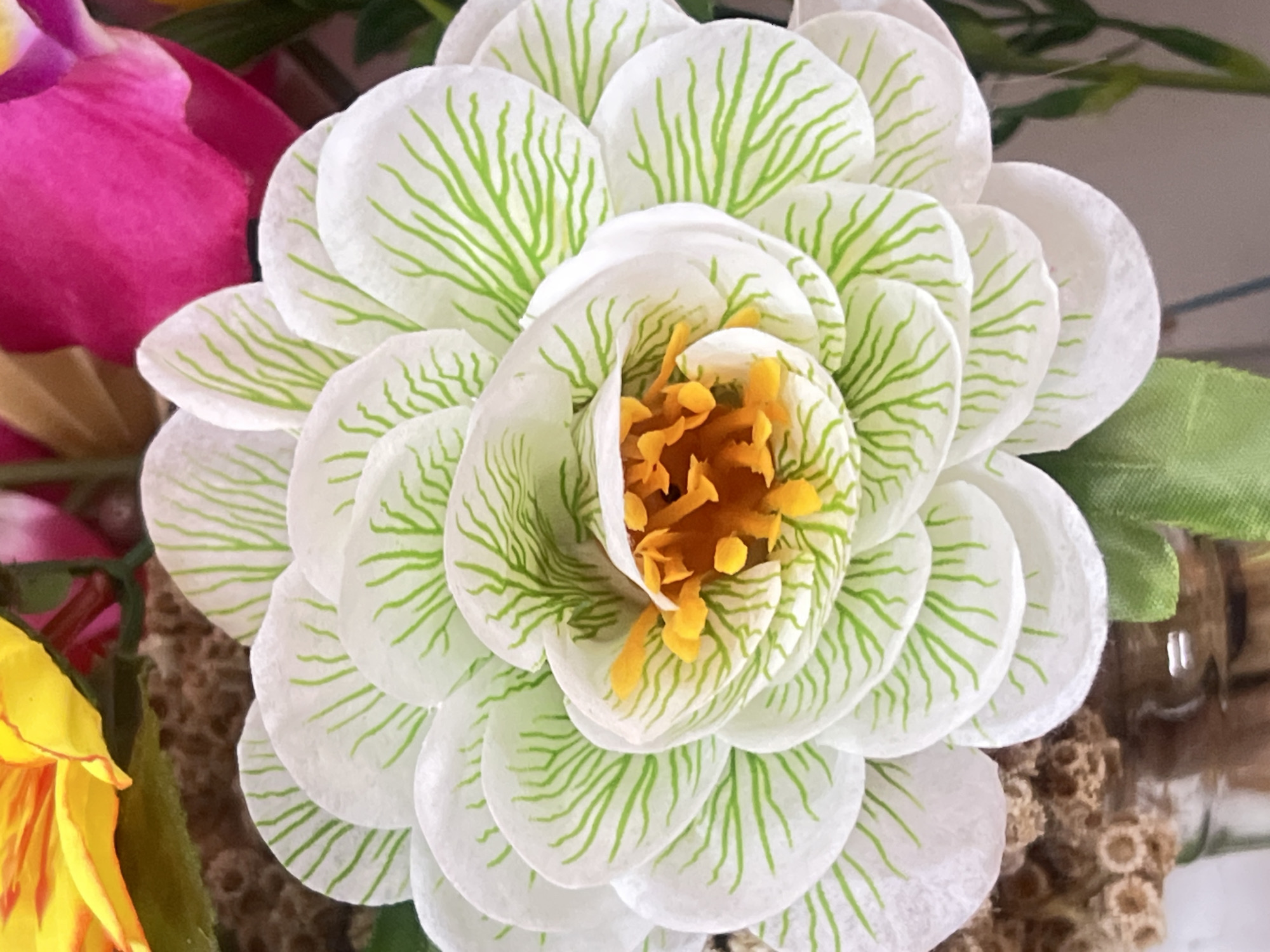 A close-up, eye-level shot shows a white, artificial flower with green veins on its petals. The flower has a bright orange center composed of stamen-like structures. Behind the white flower, portions of a yellow and a pink artificial flower are visible. The background is blurred, with hints of greenery and a textured surface that could be part of a floral arrangement or bouquet. The scene appears to be indoors, possibly in a display or home environment. There are no people present. The overall impression is of a decorative floral arrangement.