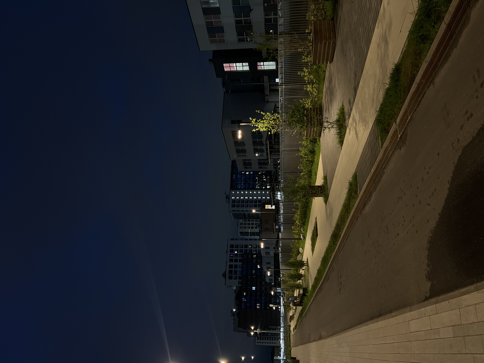 The image depicts a modern, urban nighttime scene in an area that appears to be a residential or mixed-use development. The sky is a deep, dark blue, characteristic of twilight or night. Several multi-story buildings with illuminated windows are visible in the background, suggesting an active community. The architecture appears contemporary, with clean lines and balconies.
In the foreground, a paved road runs alongside a pedestrian walkway. The road surface shows signs of wetness, with dark patches indicating moisture, possibly from recent rain or dew. The walkway is constructed with light-colored paving stones. Adjacent to the walkway is a strip of green grass and then a low metal fence. Beyond the fence, there are trees planted in wooden planters, adding a touch of nature to the urban landscape. Streetlights are also present, casting a warm glow on the scene and illuminating the pathway. The overall impression is one of a well-maintained and developing urban environment at night. No people are visible in the image.