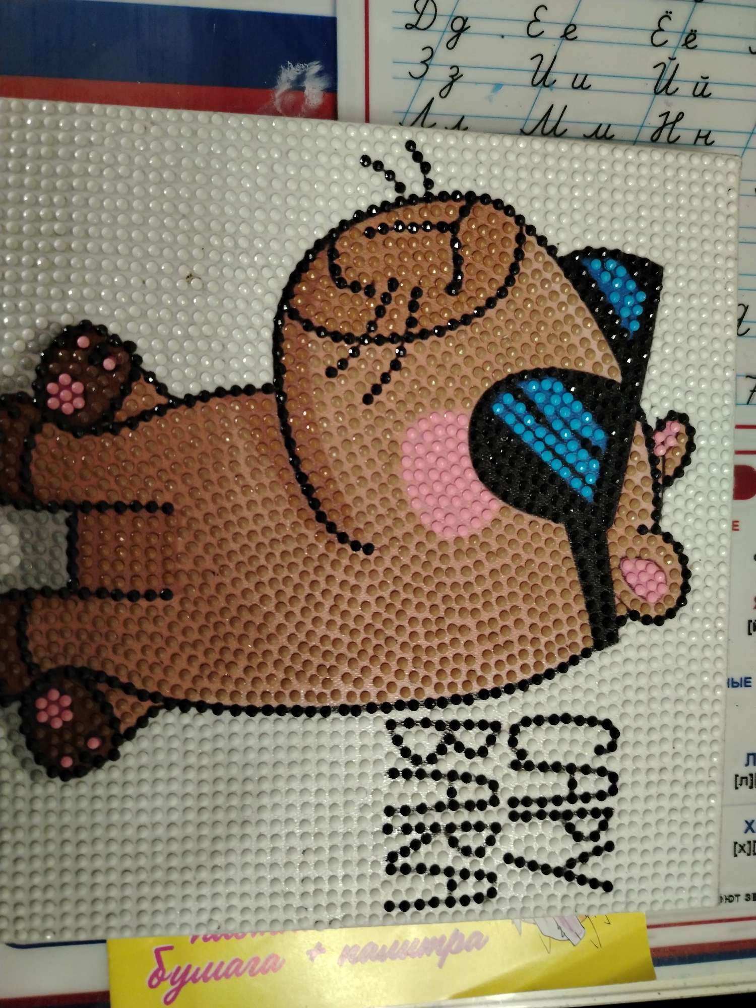 The image shows a diamond painting of a brown bear wearing sunglasses, laying on a white dotted background. The bear's head is on the right side of the image, and its body extends to the left. The bear has a pink cheek and dark brown paws. The sunglasses are black with blue gems. Below the bear, there is the text "BEAR CAPY" written in black gems. The background is white with small, uniform dots. The top of the image shows a notebook with cursive writing in an alphabet-like pattern. The bottom of the image shows a yellow paper with Cyrillic lettering that says "Бумага + каметра". There is no location given.