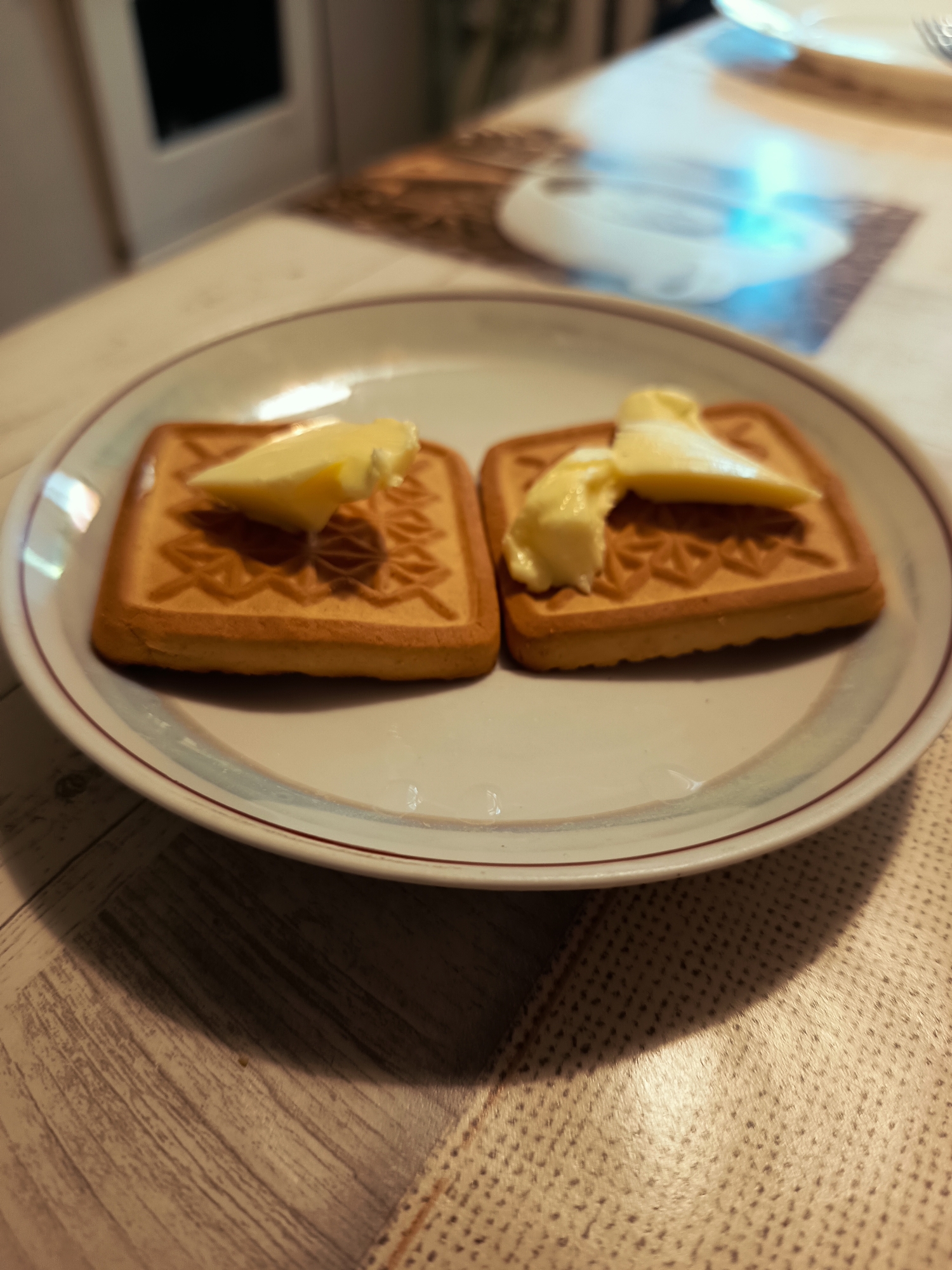 Two square biscuits with embossed patterns are on a white plate with a thin red rim. Each biscuit has a dollop of yellow butter on top. The plate is placed on a wooden table. In the background, out of focus, is a blurred image of a white and blue pattern on a placemat, and a hint of a kitchen cabinet. The scene is indoors, likely in a domestic setting. There are no people visible in the image. The image is taken from a high angle, looking down at the plate.