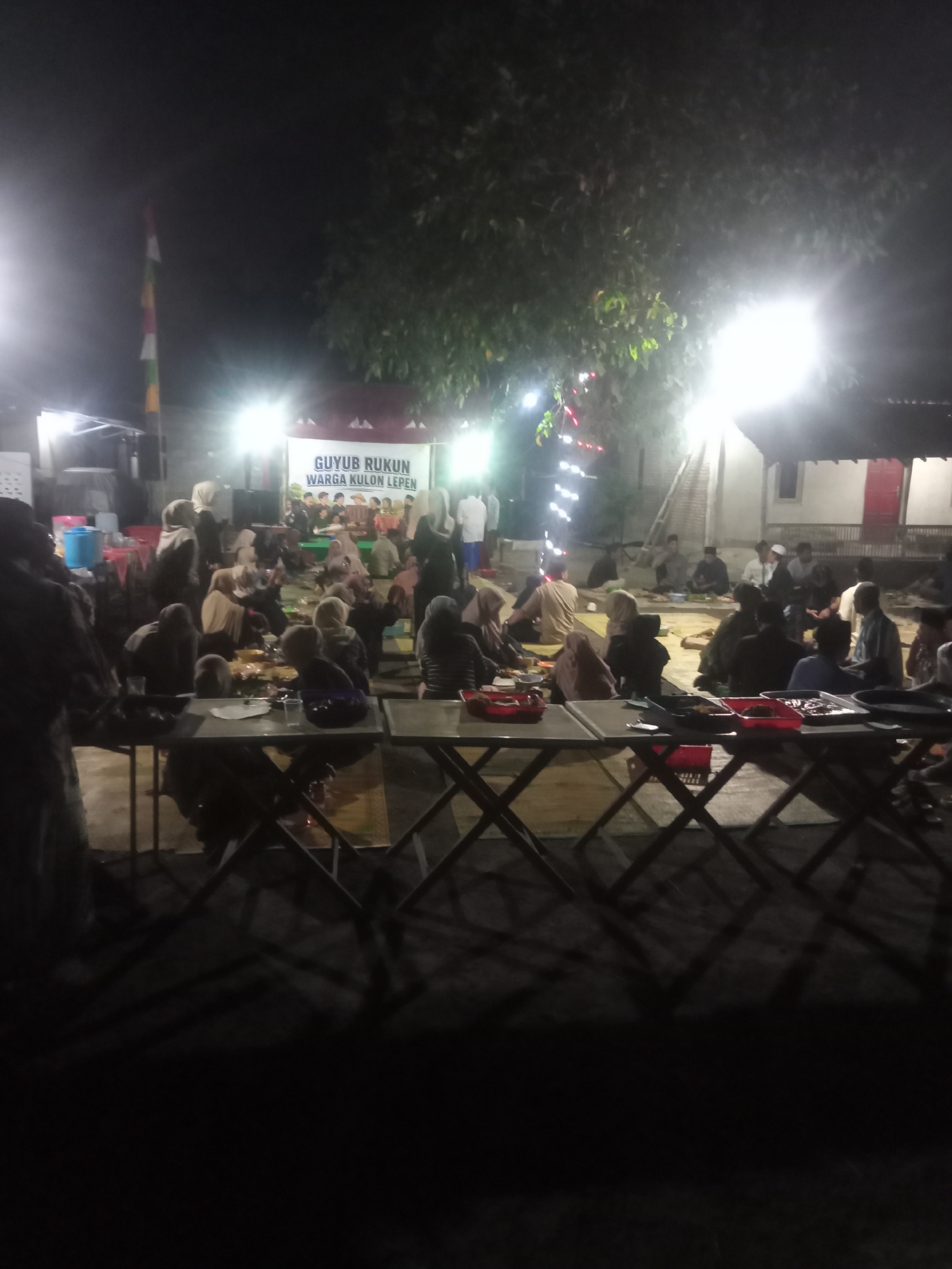 The image depicts an outdoor nighttime gathering in Jetis, Indonesia, likely a community event. A banner in the background reads "GUYUB RUKUN WARGA KULON LEPEN," suggesting a neighborhood assembly or communal activity. Numerous individuals, predominantly dressed in Islamic attire, are seated on the ground and on mats, engaged in eating and conversing. Tables are set up in the foreground with food items in trays. Decorative lights are visible in the background, strung across trees. The overall atmosphere appears to be one of social gathering and shared mealtime.