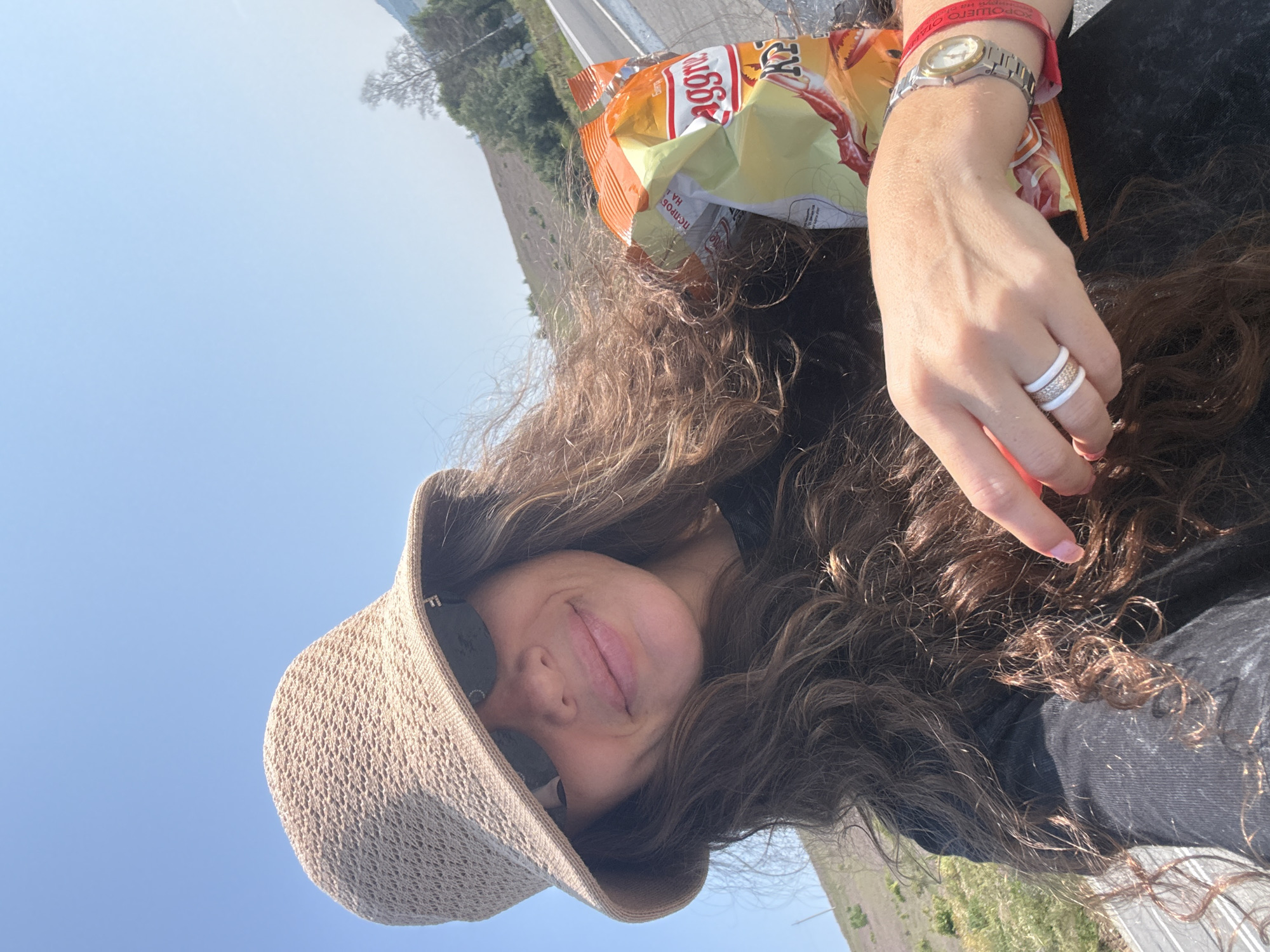 The image shows a woman wearing a straw hat and sunglasses, holding a bag of chips. She is smiling at the camera. The background is a bright blue sky and a hilly landscape with trees and a road. The location is identified as Choya, Russia.