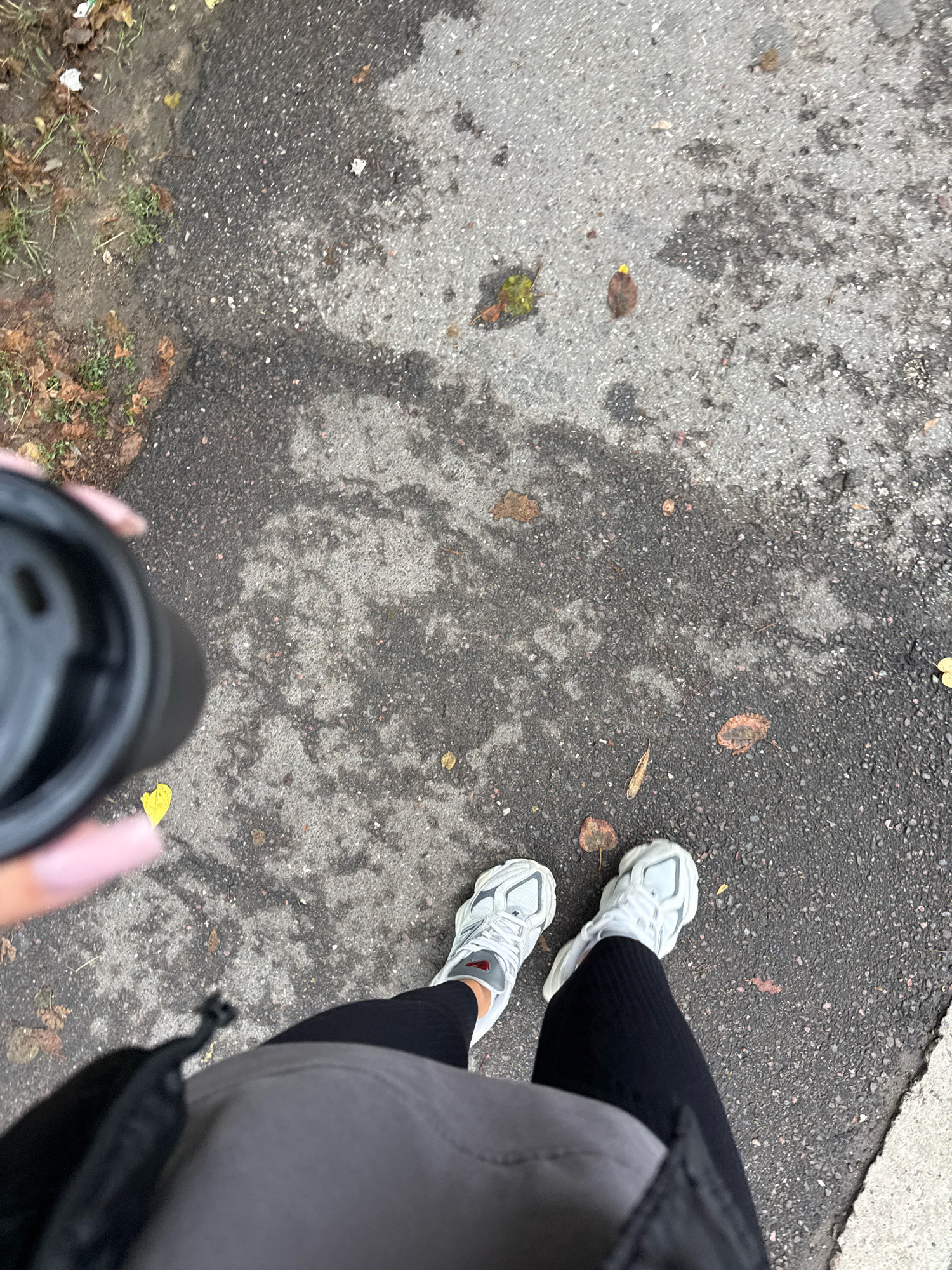 A high-angle shot looks down at a person's feet, clad in white athletic shoes and black leggings, standing on a wet asphalt path in Chișinău, Moldova. A black disposable coffee cup is held in the person's left hand, which is partially visible at the top left of the frame. Fallen leaves are scattered across the path, indicating an autumnal setting. The path is bordered by a strip of grass with more scattered leaves to the left and a lighter colored paved area to the right. The overall scene is outdoors, with an emphasis on the textures of the ground and the casual attire of the individual.