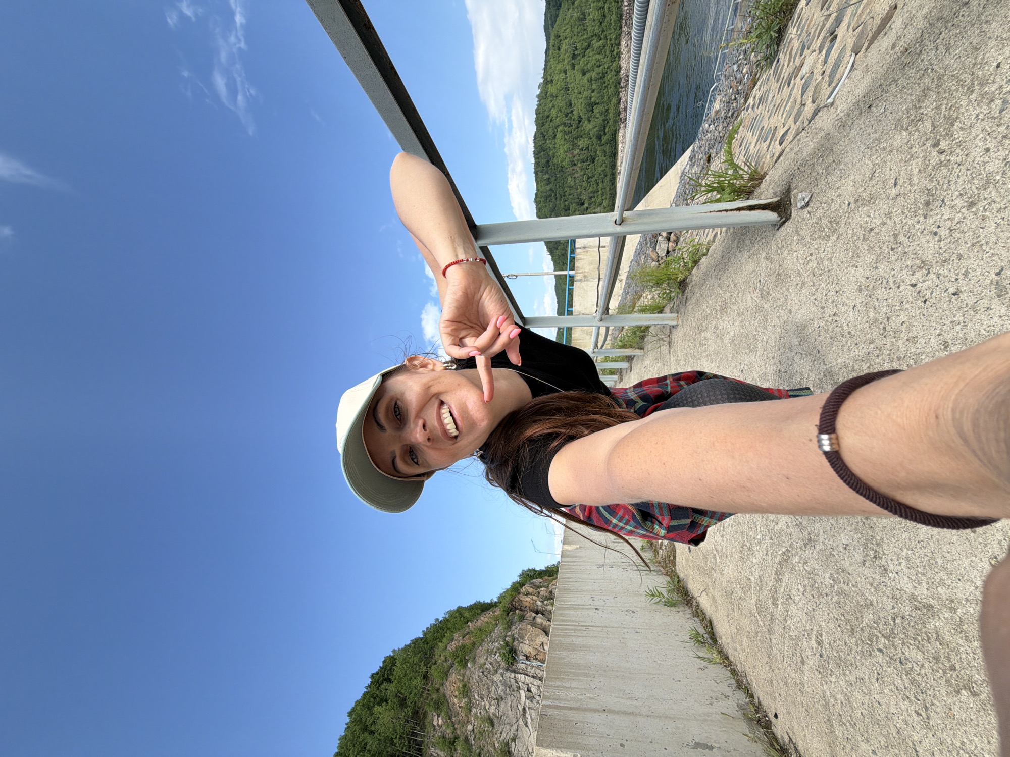 A woman is taking a selfie on a paved pathway next to a metal railing. She is wearing a black t-shirt, a red and black plaid shirt, a light green baseball cap, and a red bracelet on her left wrist. She has long dark hair and is smiling at the camera, with her right hand raised and her index finger and thumb touching. The pathway appears to be on a dam or a similar structure, with a body of water and forested hills in the background. The sky is blue with a few scattered clouds. The location is the Talakan Dam in Talakan, Russia.