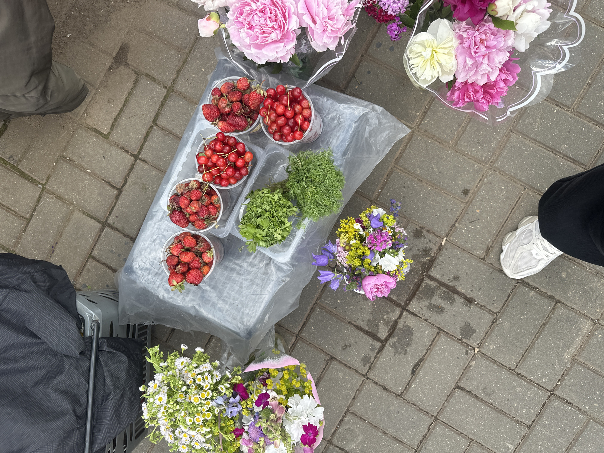 The image captures an overhead view of a street market stall displaying fresh produce and bouquets of flowers. The primary subjects are two buckets of strawberries, two buckets of cherries, and two containers of leafy greens, likely herbs. Several bouquets of flowers, including pink peonies, white flowers, and smaller colorful blooms, are arranged around the produce. The setting is outdoors on a paved sidewalk made of grey rectangular bricks.
The activity appears to be a sale at a market, with a person's legs and feet visible on the left and right sides of the image, suggesting they are either the vendor or a customer. The person on the left is wearing a dark grey jacket and standing near a dark grey shopping bag. The person on the right is wearing black pants and white sneakers.
Notable details include the arrangement of the produce in clear plastic cups and containers, suggesting they are for sale by weight or portion. The flowers are also presented in bouquets, some wrapped in clear plastic. The lighting suggests it is daytime, and the cobblestone ground appears slightly damp in places, hinting at recent rain or mist. There is no visible text in the image. The overall impression is one of a vibrant, fresh market day.