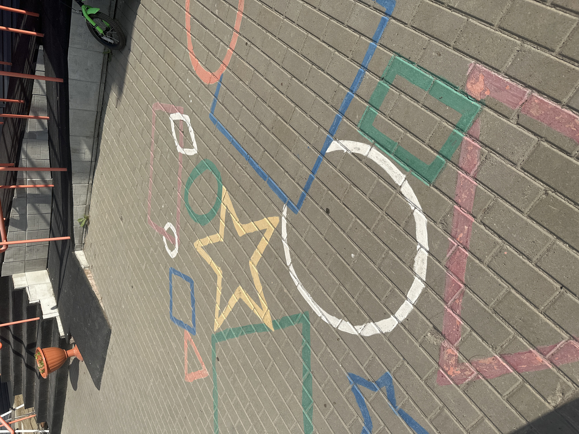A close-up, high-angle shot shows chalk drawings on a paved outdoor surface, likely in Novoaltaysk, Russia. The drawings depict a hopscotch grid with various geometric shapes including squares, circles, and a star, rendered in multiple colors such as pink, blue, yellow, green, and white. The paving stones are arranged in a diagonal pattern. In the background, a grey concrete wall and a black ramp leading to stairs are visible, along with an orange planter with a plant. A green scooter is parked to the right. The scene is illuminated by sunlight, casting shadows.