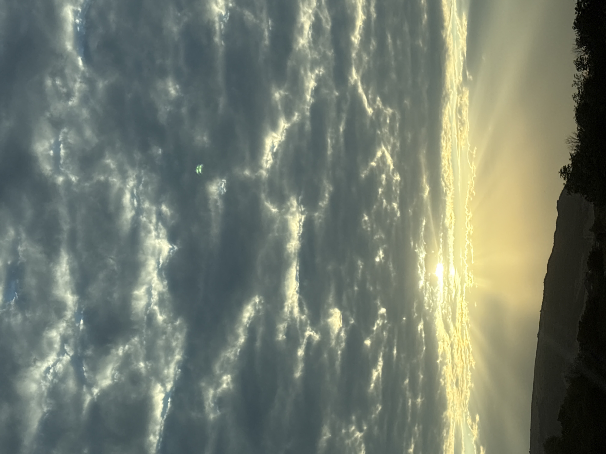 The image captures a dramatic sky over Lysogorskaya, Russia, likely at sunrise or sunset. Dark, textured clouds dominate the upper half of the frame, with streaks of sunlight piercing through, creating a volumetric effect. The sun itself is partially obscured by a bank of clouds near the horizon, casting golden rays that illuminate the sky. Below the clouds, a silhouetted landscape of hills and trees stretches across the bottom of the image. In the sky, a small, bright green object is visible, its nature and purpose unclear, but appearing as a distinct point of color against the overcast sky.