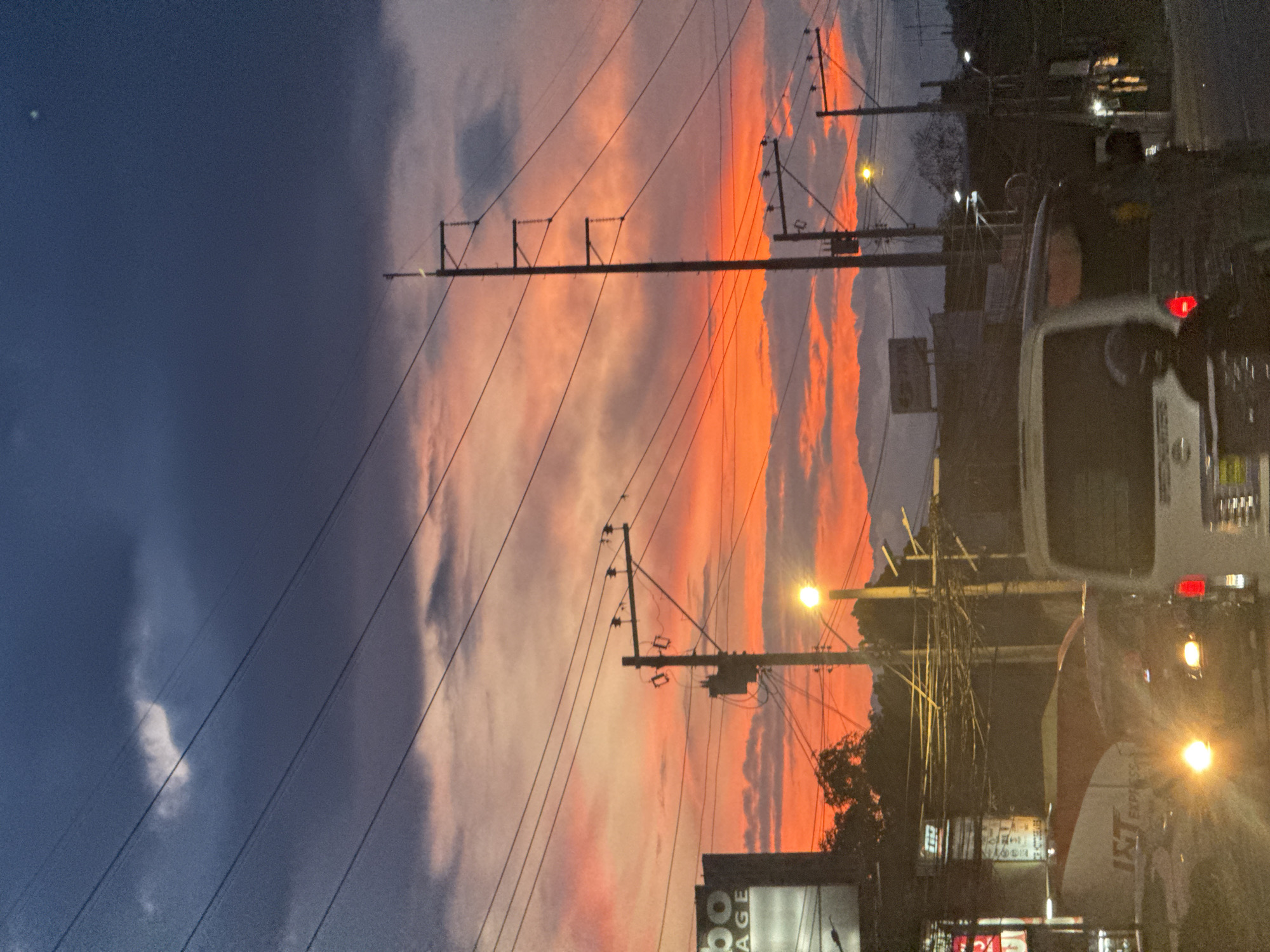 The image shows an outdoor scene at dusk in Davao City, Philippines. Overhead power lines and utility poles are silhouetted against a sky streaked with orange and gray clouds. Streetlights cast a glow on nearby buildings and vehicles, which include a white van with visible taillights. The scene depicts a busy street with commercial establishments on either side. The sky suggests the sunset, and the foreground shows signs of urban infrastructure and activity.