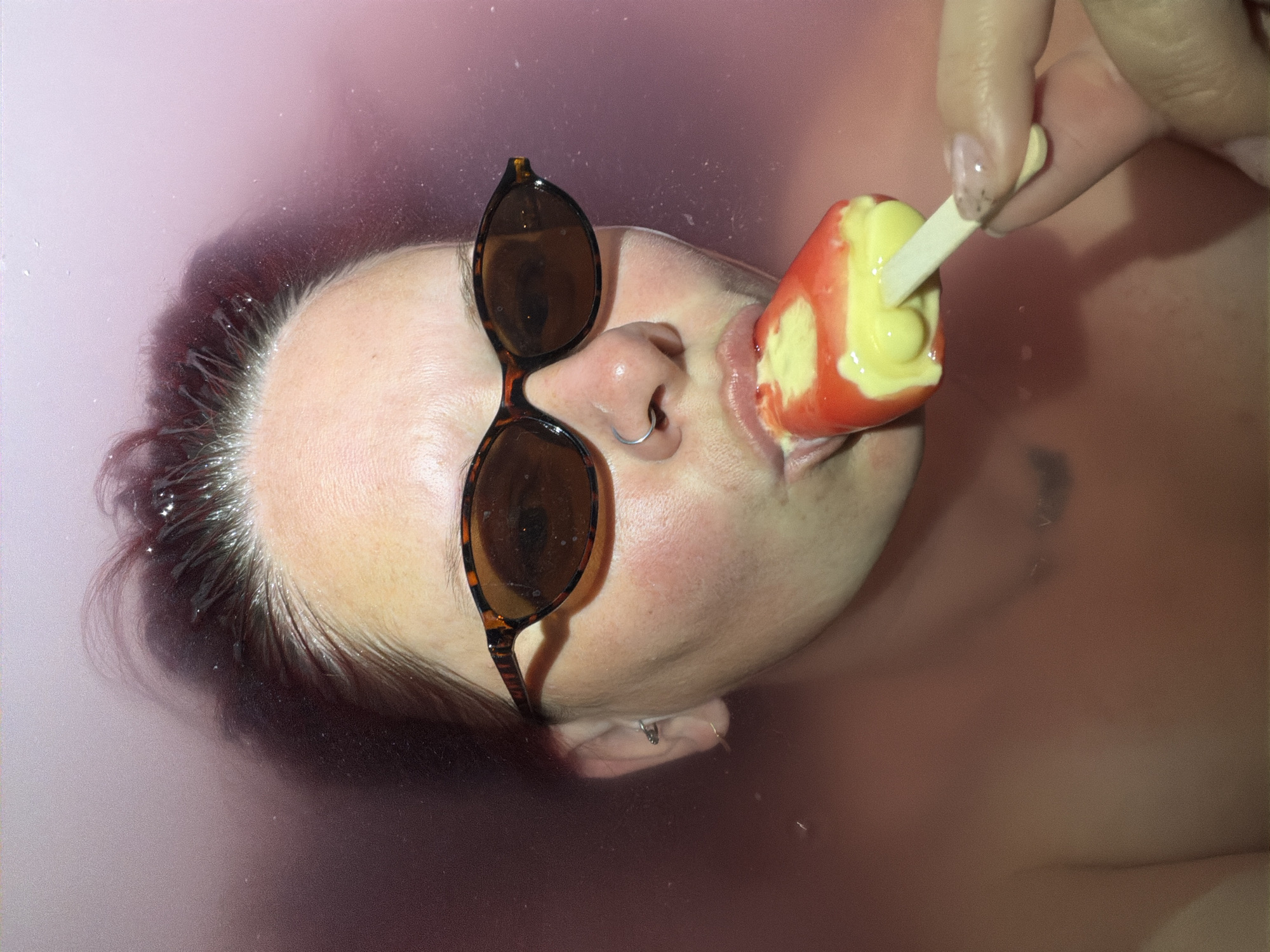 A medium close-up shot depicts a person submerged in water, likely in a bathtub, with a pinkish hue to the water. The person is wearing tortoiseshell sunglasses perched on their nose and has a silver nose ring. They are in the process of eating a red and yellow popsicle held by a hand with painted fingernails. The individual’s hair appears to be wet, and a faint tattoo is visible on the chest or upper torso. The location is indeterminate.