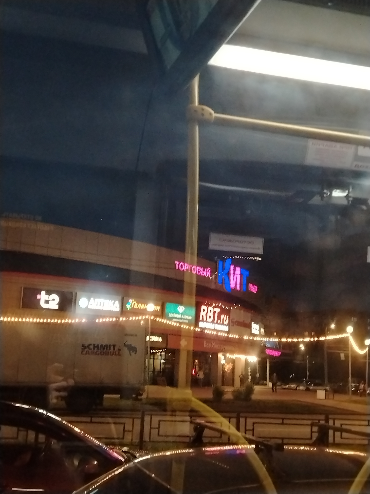 The image presents a nighttime street scene in Izhevsk, Russia, captured from inside a vehicle, likely a bus, evidenced by the prominent yellow handrail and interior reflections on the window.
The central subject is a brightly illuminated commercial building, its facade decorated with numerous warm-toned string lights. Several commercial signs are visible: a large neon sign at the top reads "ТОРГОВЫЙ КИТ" (TORGOVY KIT), indicating a shopping center. Below it, a red sign displays "RBT.ru," an electronics store. To its right, a partial sign says "Все Инстру" (likely part of "Все Инструменты," a hardware store). Further left, other visible signs include "t2," "АПТЕКА" (APTEKA – Pharmacy), and "Галам" (likely "Галамарт," a household goods store). A green sign for "ЗЕЛЕНЫЙ КАМЕНЬ" (ZELENY KAMEN – Green Stone) is also seen.
On the street level, a large white truck is parked, displaying the brand "SCHMITT CARGOBULL" along with an image of a bull. The tops of other cars are visible in the foreground, reflecting the interior lights and structure of the vehicle from which the photo was taken. A few pedestrians are discernible on the sidewalk, walking past the illuminated storefronts. The scene is characterized by the glow of artificial lights against the dark night sky, typical of an urban commercial district after sunset.