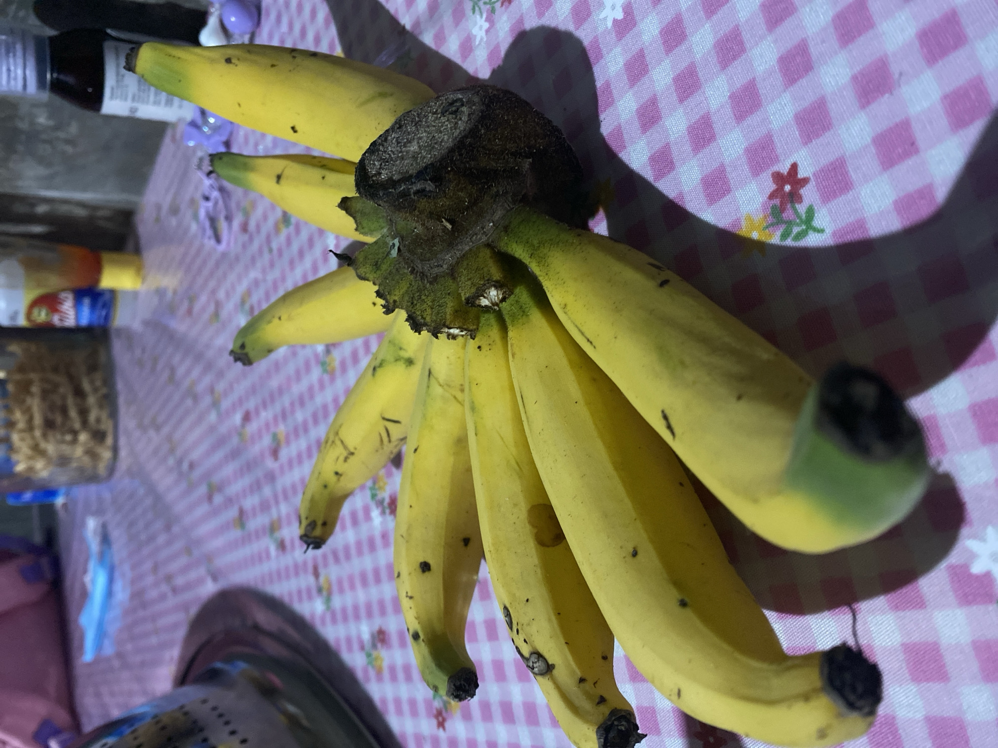 In Bontoharu, Indonesia, a bunch of ripe bananas is arranged on a pink and white checkered tablecloth. The bananas are predominantly yellow with some brown spots, indicating ripeness. The stems of the bananas are dark and clustered together. The tablecloth has a subtle floral pattern. In the background, out of focus, are various objects including bottles, a container of food, and a blue mask, suggesting a domestic setting. The lighting creates shadows from the bananas on the tablecloth.