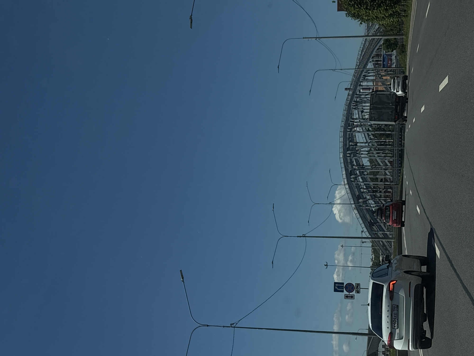 This is a daytime, eye-level, horizontal shot of a highway in Saint Petersburg, Russia, with a large metal bridge in the background. The sky is clear and blue. On the highway, there are several vehicles. In the foreground, a white Skoda Octavia is driving in the right lane. Behind it, a red hatchback is in the left lane. Further ahead, a large truck is in the right lane, followed by a white SUV in the left lane. Along the side of the highway, there are tall streetlights. The bridge is a complex metal structure with a series of arches. Trees and some buildings are visible behind the bridge. Traffic signs are present on the side of the road.