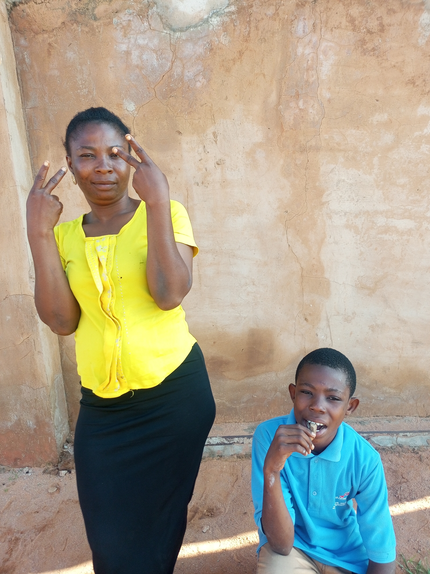 A medium shot captures two individuals posing against a textured, light brown wall. On the left, a woman stands with her left arm raised, fingers forming a 'V' sign towards the camera, and her right hand also displaying a 'V' sign. She wears a vibrant yellow collared shirt and a long black skirt. Her expression is direct, and she has dark hair pulled back.
To the right and slightly lower, a young boy is seated, looking towards the camera with his mouth open and holding a small object in his hand, possibly food, near his mouth. He is wearing a light blue collared shirt. The wall behind them is a rough, weathered surface with visible cracks and imperfections. The ground appears to be sandy or dirt. The lighting suggests it is daytime, with shadows indicating sunlight. The overall scene is simple, with the focus on the individuals and their interaction with the camera. The location is identified as Tempure, Nigeria.