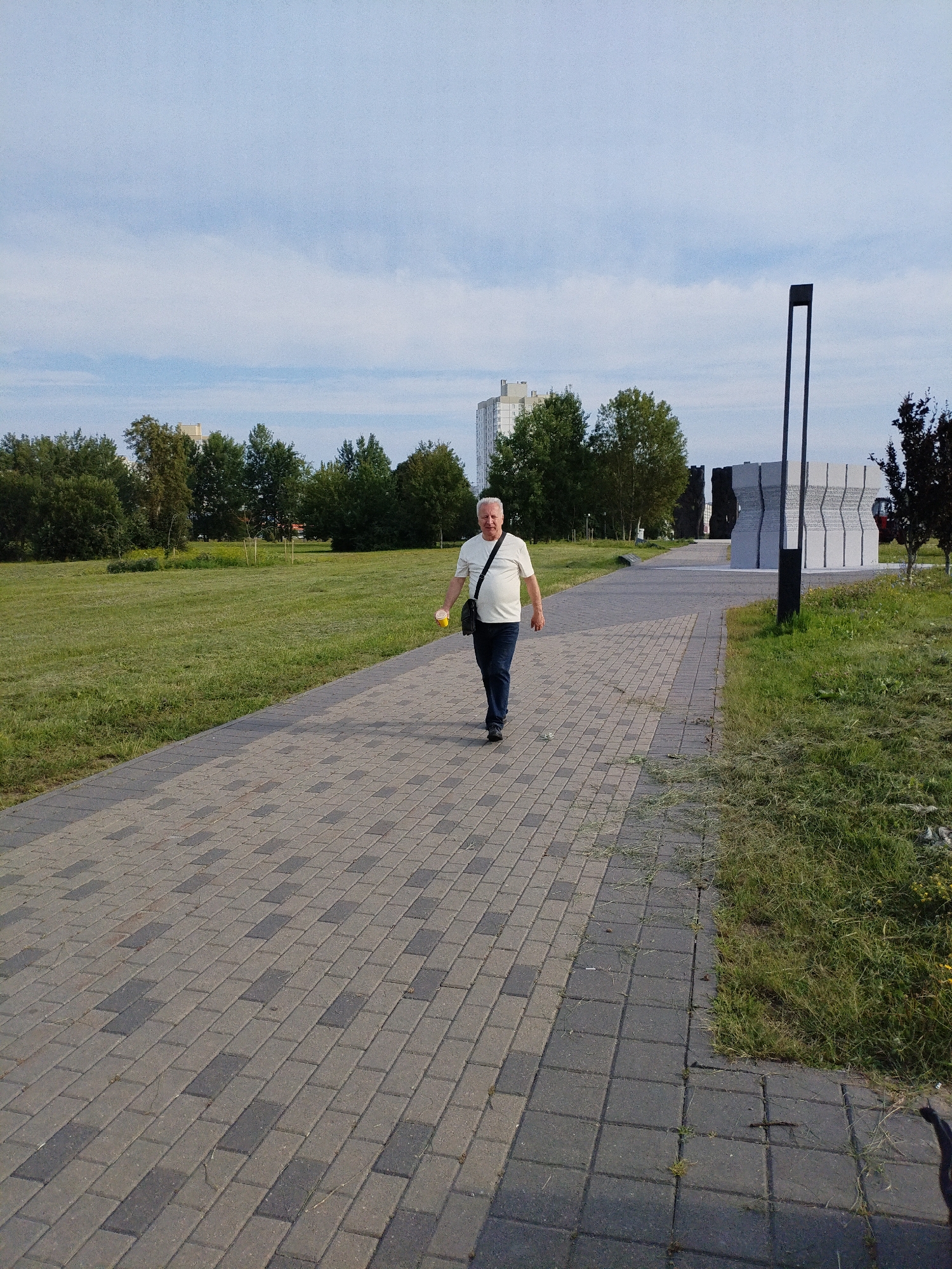 A man is walking on a paved walkway in a park in Minsk, Belarus. The man is wearing a white t-shirt, blue jeans, and black shoes. He is carrying a black bag over his left shoulder and a yellow cup in his right hand. The walkway is made of gray and dark gray pavers. To the left of the walkway is a grassy area with trees in the background. To the right of the walkway is a modern sculpture and a tall, dark pole with a light fixture. In the distance, there are several tall buildings. The sky is blue with some white clouds.