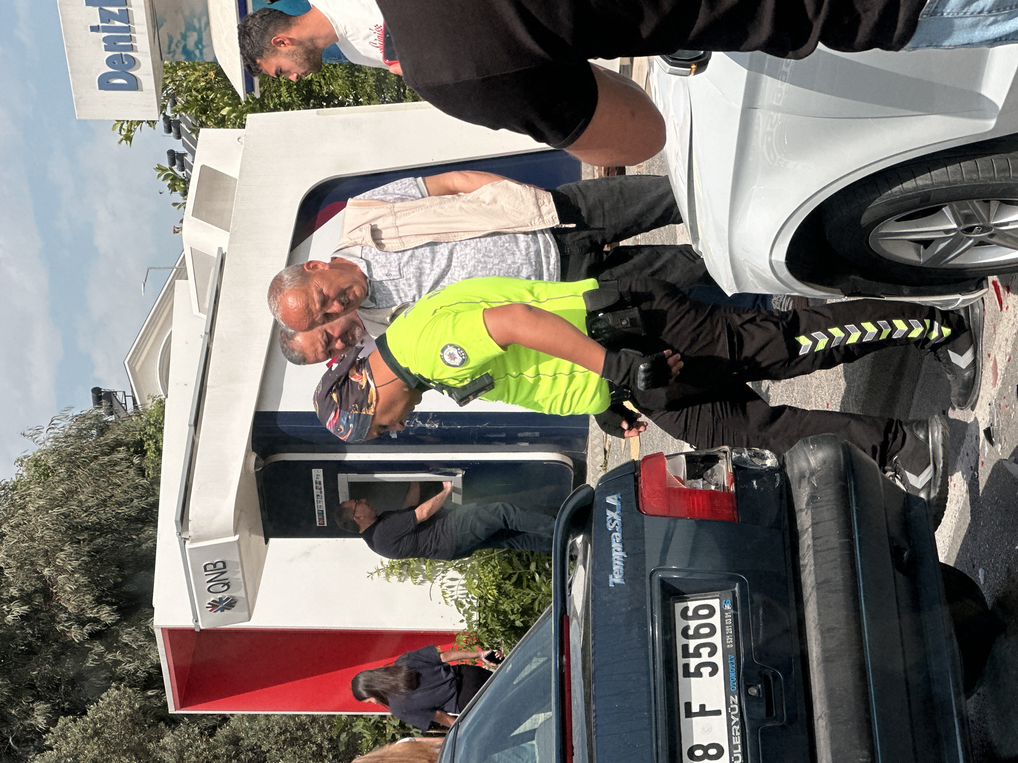 The image shows a street scene in Fethiye, Turkey. A QNB Bank ATM is visible, with a man using it. A police officer in a bright green vest stands nearby, seemingly monitoring the situation, along with two other men. A Denizl Bank sign is partially visible in the background. A dark blue car with a damaged tail light is parked, with a license plate that reads "8 F 5566". "Tempra SX A" is written on the car's trunk. A white car is parked on the opposite side. The surrounding environment includes trees and other people.