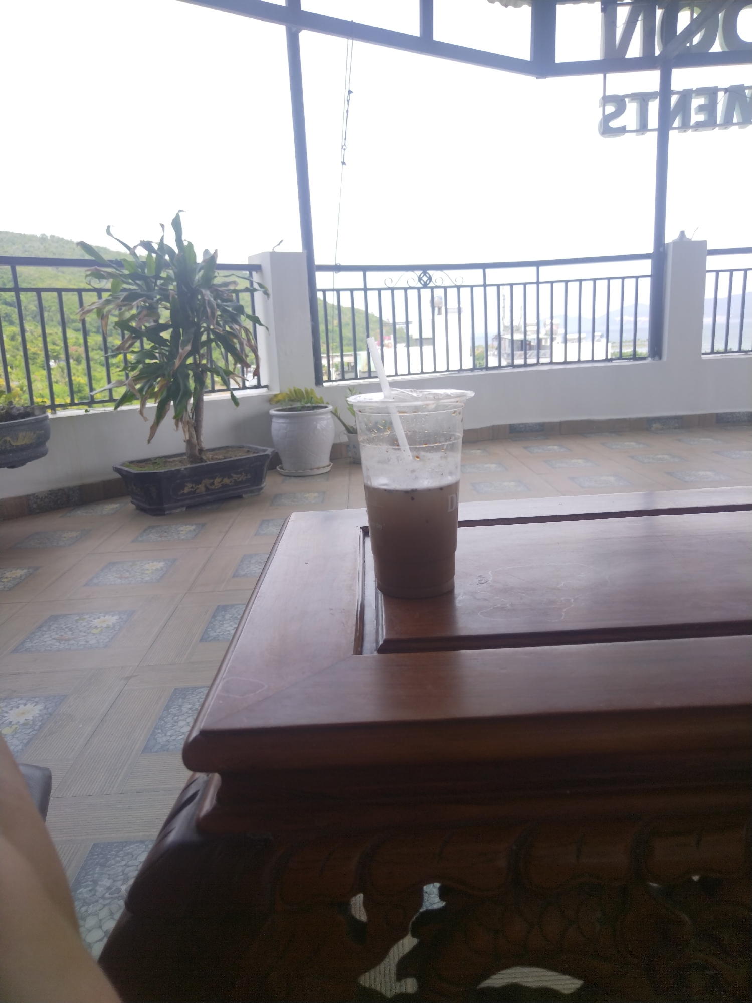 An iced coffee sits on a wooden table on a balcony overlooking a lush green hillside and the ocean in Bac Nha Trang Ward, Vietnam. The balcony is tiled with a patterned design. In the background, a black metal railing separates the balcony from the hillside. A tall, leafy plant in a rectangular planter is visible to the left of the coffee. The sky is overcast. The coffee cup is clear plastic with a straw.