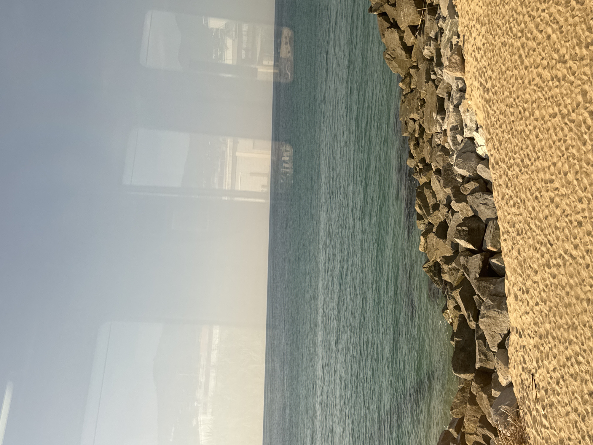 The image depicts a coastal scene in Cabrera de Mar, Spain, viewed through a window. The foreground features a sandy beach with footprints and scattered dried vegetation. Behind the beach, a rocky breakwater separates the sand from the sea. The sea itself is a vibrant turquoise color, with gentle waves. In the distance, the hazy horizon shows buildings and mountains, possibly part of the town or surrounding landscape. Reflections of a window frame and possibly interior elements are visible on the surface of the water and in the sky, suggesting the photograph was taken from inside a moving vehicle or building. The sky is a pale blue, with some atmospheric haze.
