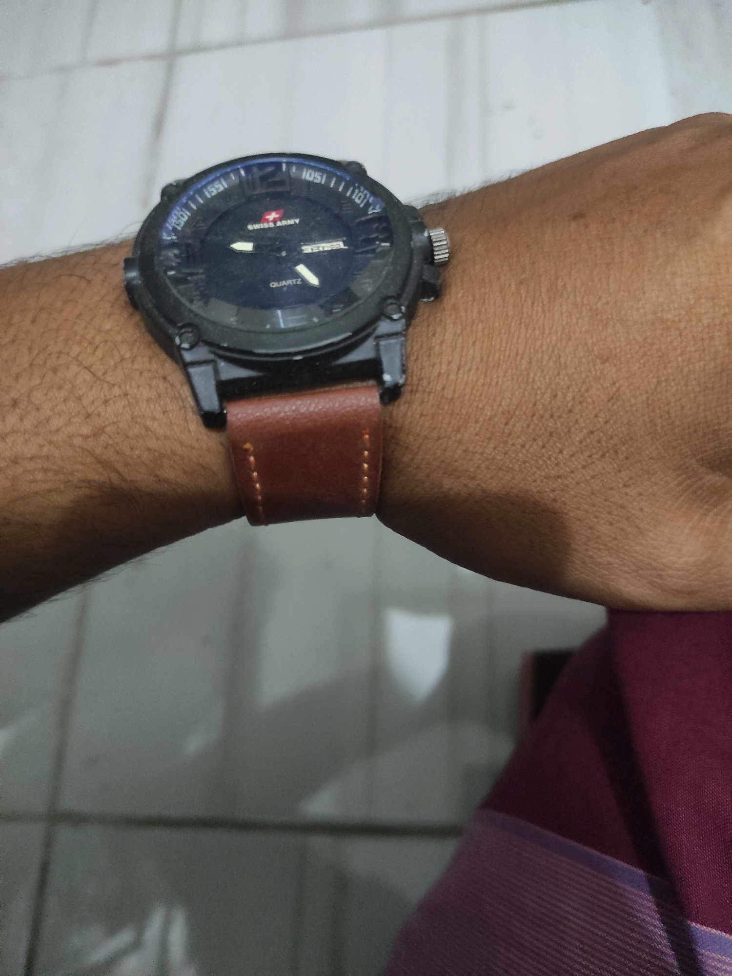 In Buntalan, Indonesia, a person's arm, adorned with a black Swiss Army watch with a brown leather strap, is visible. The watch face features a red Swiss Army logo and is marked with "QUARTZ". The background consists of a tiled floor with grid lines and a shadow. The person is wearing a maroon garment.