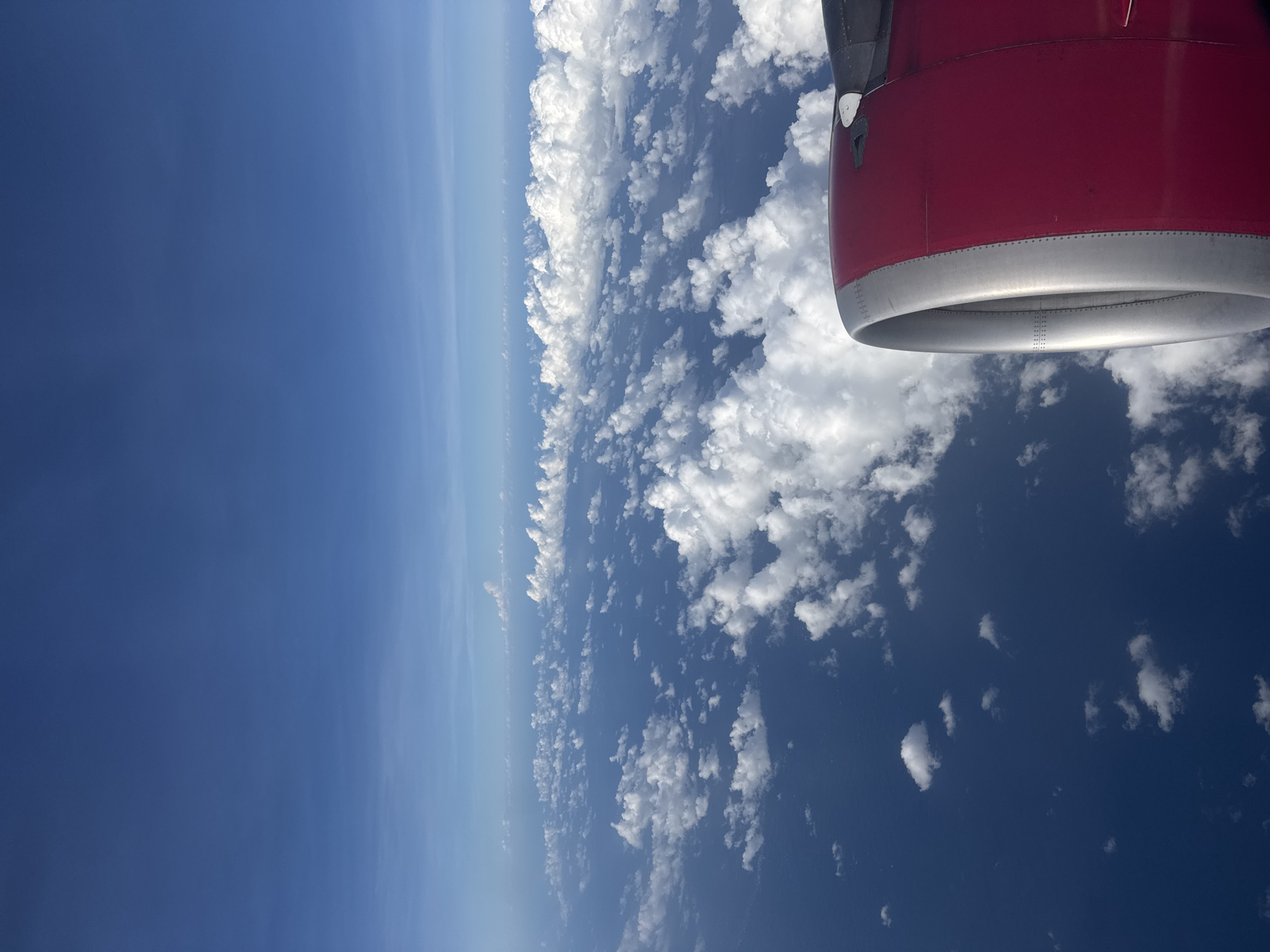 The image is an aerial view from an airplane window. A portion of the airplane's red and silver engine is visible in the upper right corner. The view outside shows a clear blue sky meeting a dense layer of white clouds. Below the clouds, patches of dark blue water are visible.