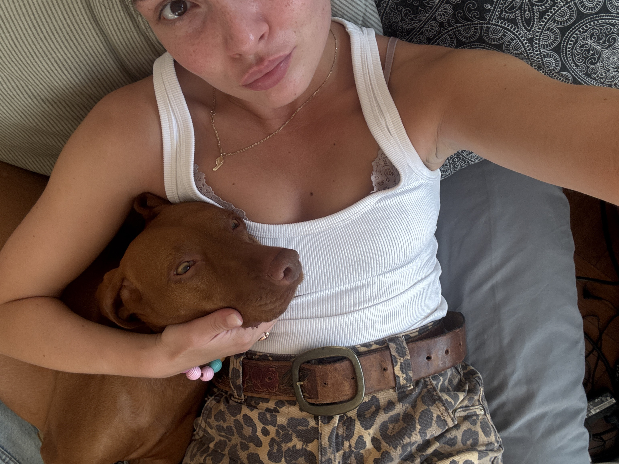 This overhead selfie captures a person and their dog in a cozy indoor setting in Sofia, Bulgaria. The central subjects are a person, likely female, and a reddish-brown, short-haired dog, possibly a Vizsla or similar breed, resting comfortably together.
The person is lying down, looking towards the camera with a relaxed expression and slightly pursed lips. She has freckles on her face and dark eyes. She is wearing a white ribbed tank top, featuring a delicate lace trim along the neckline. A thin gold necklace with a small charm, possibly a shoe or wing, is visible around her neck. Her lower body is clad in leopard-print pants or shorts, cinched with a wide, distressed brown leather belt featuring a large, ornate metal buckle. Around the dog's neck, or perhaps on the person's wrist close to the dog, is a colorful beaded bracelet with pink, light blue, and white beads.
The dog lies with its head resting on the person's arm and chest, appearing calm and comfortable, gazing gently towards the side. Its fur is a consistent reddish-brown color.
The background suggests an indoor environment, likely a bedroom or living space. To the left, a light-colored striped pillow is visible, while to the right, a darker gray or blue solid fabric, possibly a blanket or pillow, can be seen. A dark fabric with an intricate white pattern (perhaps a throw pillow or blanket) is also present behind the person's right arm. A hint of wooden flooring is visible on the far right.
The overall scene conveys a feeling of affection and peaceful companionship between the person and their pet. The lighting appears natural, suggesting it is daytime. There is no visible text in the image.