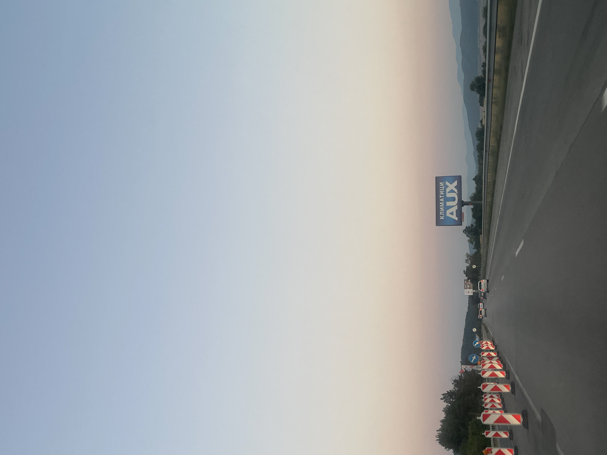 The image captures a highway scene during what appears to be dusk, with a soft gradient of blue to pink hues in the sky. The highway itself is asphalt, with visible lane markings. On the left side of the road, a series of traffic barrels, alternating red and white reflective stripes, are lined up, indicating potential road work or a lane closure. Several vehicles, including cars and what looks like a truck, are traveling on the highway. In the distance, a billboard with the word "AUX" and Cyrillic text above it, likely advertising air conditioning units, is visible. Rolling hills and mountains form the backdrop of the scene, with trees dotting the landscape. The overall environment suggests a rural or semi-rural setting on the outskirts of Ihtiman, Bulgaria.