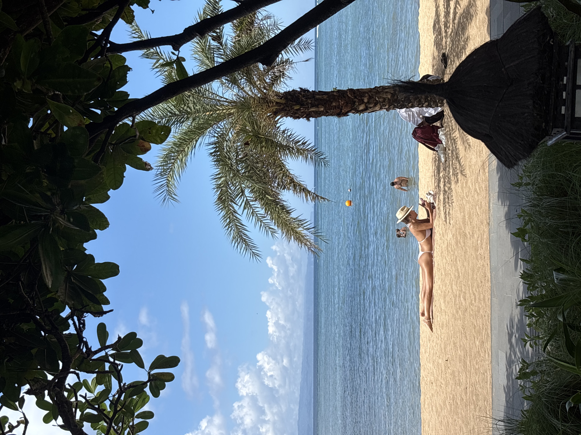 The scene is a beach in Denpasar, Indonesia. A woman wearing a white bikini and hat lies on the sand, reading a book. Nearby, a person stands in the water. A palm tree casts a shadow on the beach. There is an orange ball floating in the water. A grass umbrella is partially visible. Trees and foliage frame the edges of the image. The sky is blue with scattered clouds.