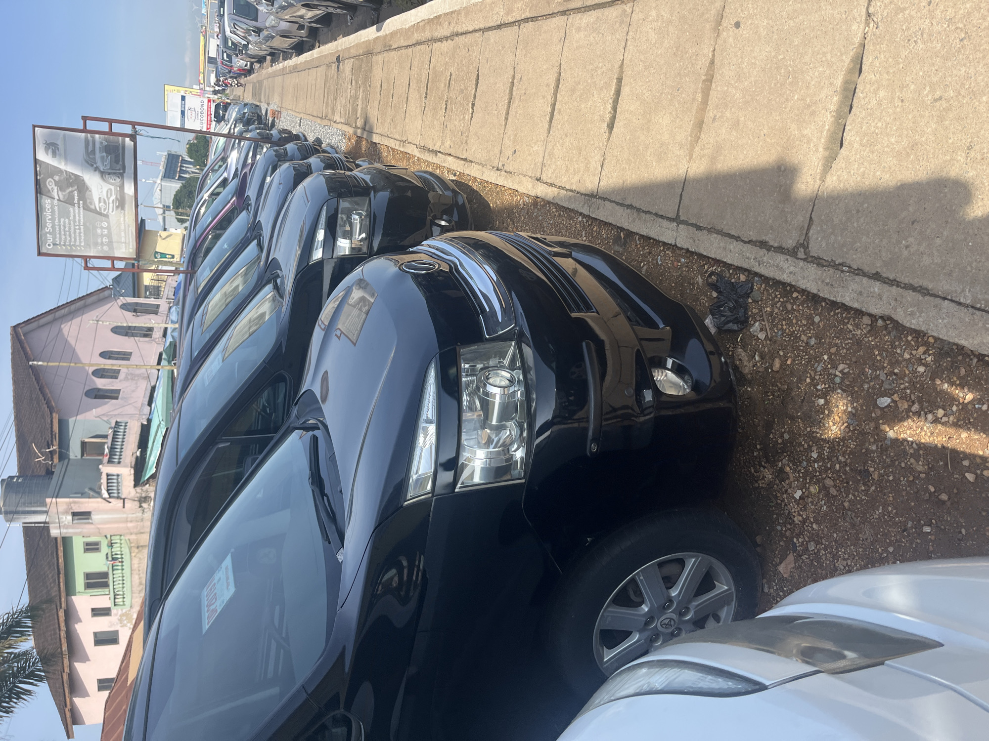 An eye-level, medium shot captures a car lot in Accra, Ghana, on a sunny day. In the foreground, a line of black vehicles, primarily minivans, are parked on a gravel lot adjacent to a concrete sidewalk. The vehicles are the primary focus, with their headlights and grills clearly visible. A white vehicle is partially visible in the lower left corner of the frame. In the background, colorful buildings with balconies stand under a clear blue sky, suggesting an urban environment. A sign detailing "Our Services" is visible on the right, with a list of automotive repair services. Other cars are parked in the distance along a street.